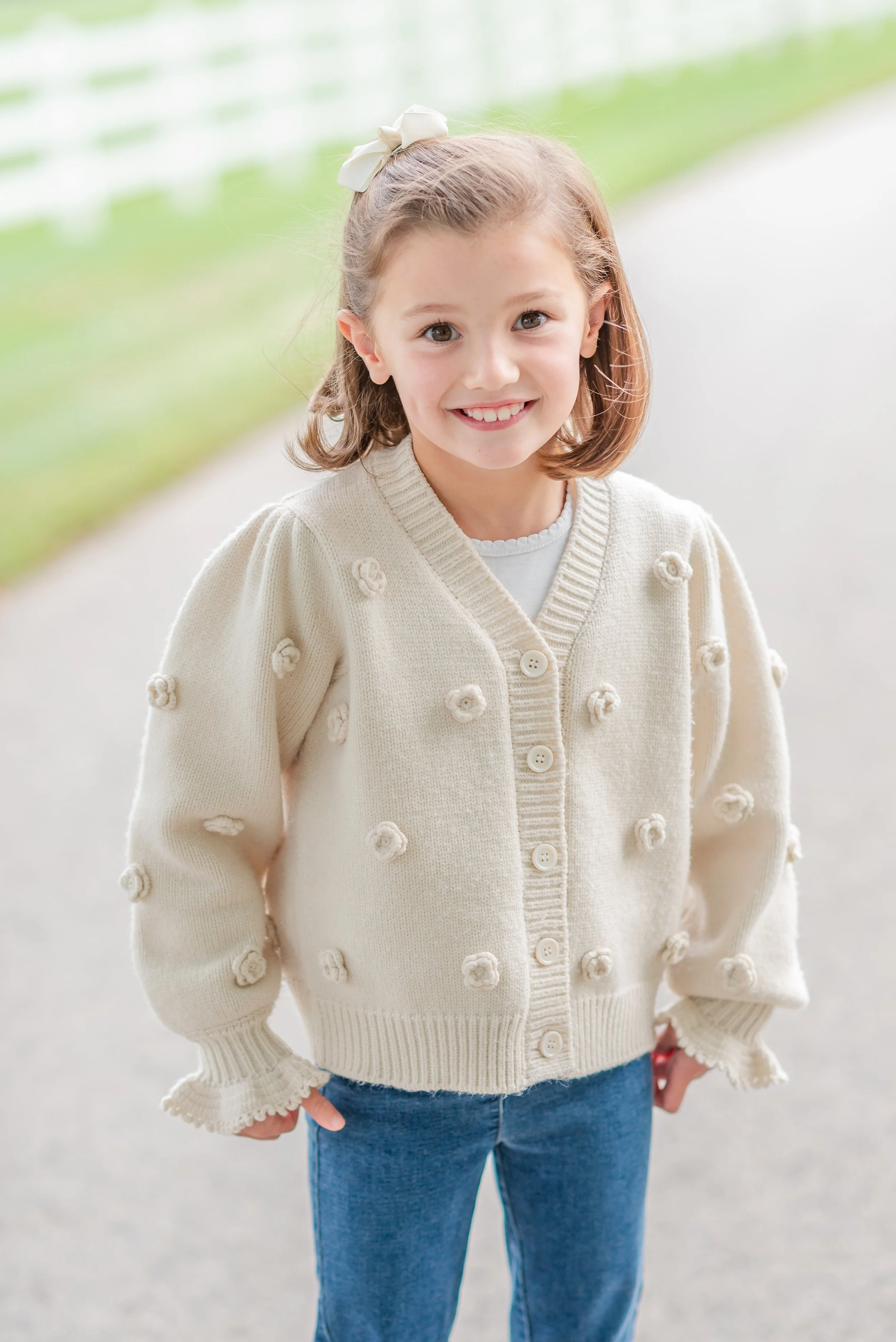 A young girl with shoulder-length light brown hair, smiling outdoors. She is wearing a cream-colored, knit cardigan with flower decorations and blue jeans.