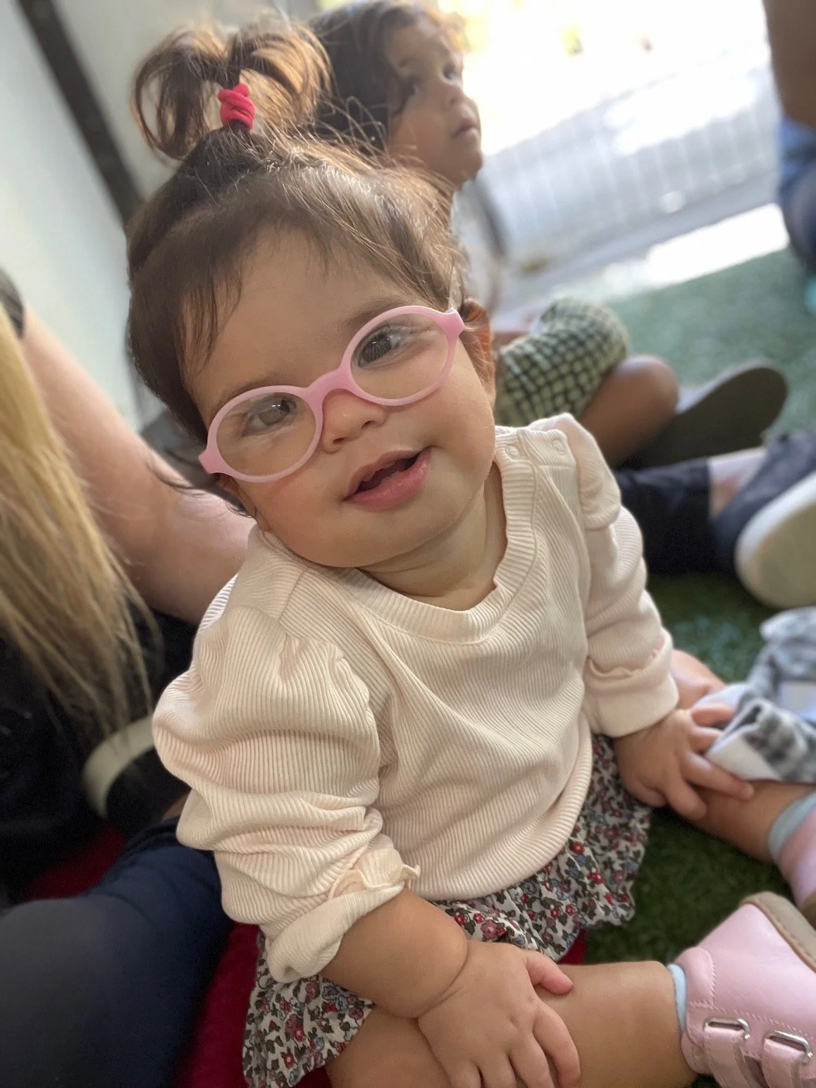 A young girl with glasses smiling and sitting on the floor, with two other children in the background.
