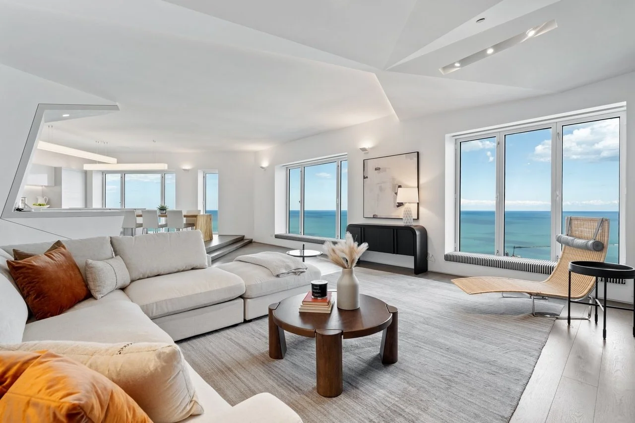Modern living room with large windows overlooking the ocean, white sofa with orange pillows, wooden coffee table with books and vase, beige lounge chair, and minimalist decor.