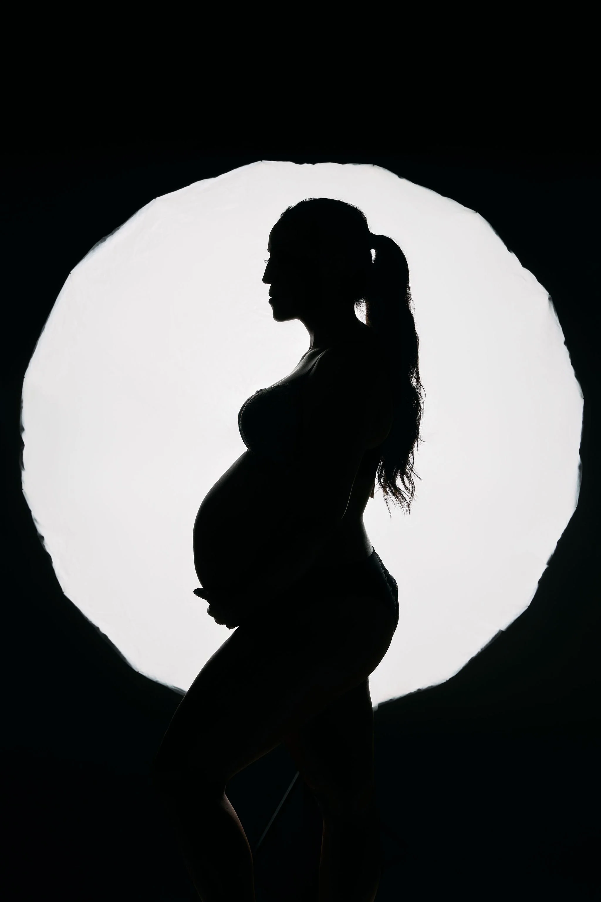 Artistic silhouette maternity portrait of a pregnant woman in profile, posed against a large circular white light.