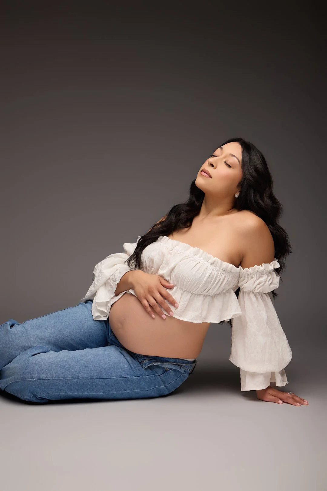Studio maternity portrait of an expectant mother seated on the floor in jeans and a white off-the-shoulder blouse, gently cradling her baby bump against a gray backdrop.