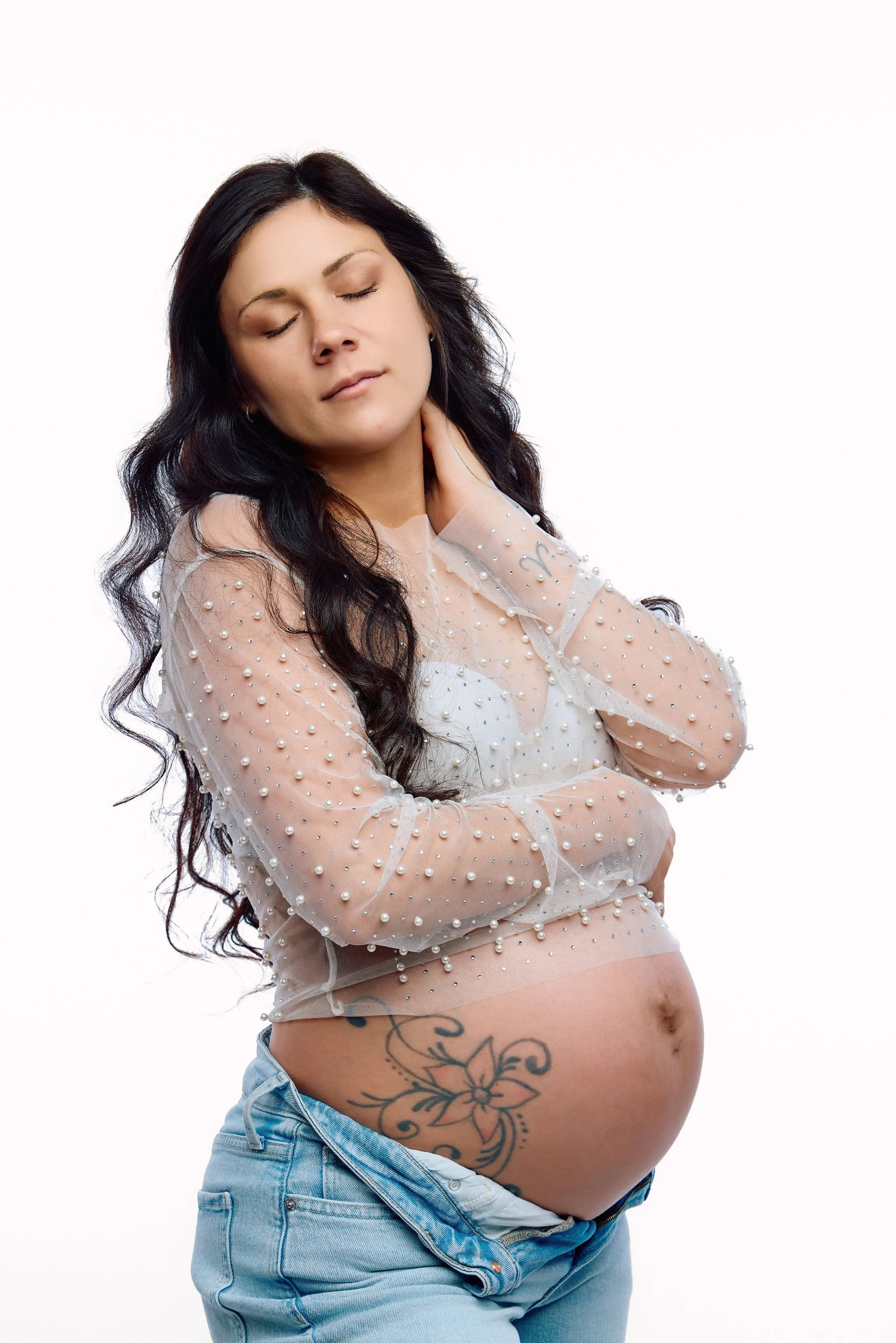 Studio maternity portrait of an expectant mother in a sheer pearl-embellished top and jeans, posing with her eyes closed and her tattooed baby bump exposed against a white background.