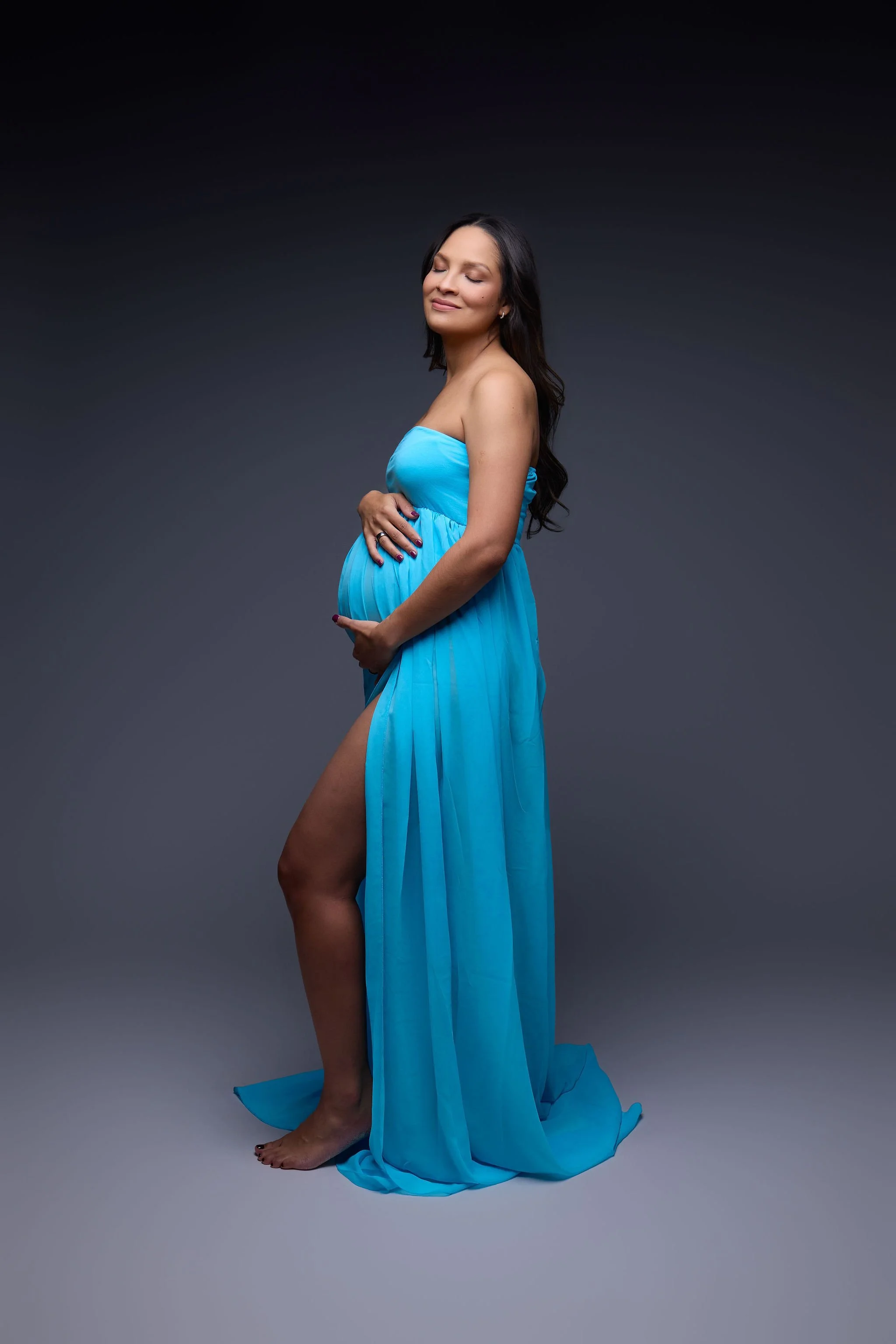 Studio maternity portrait of a pregnant woman in a flowing blue gown, cradling her belly against a dark gray background.