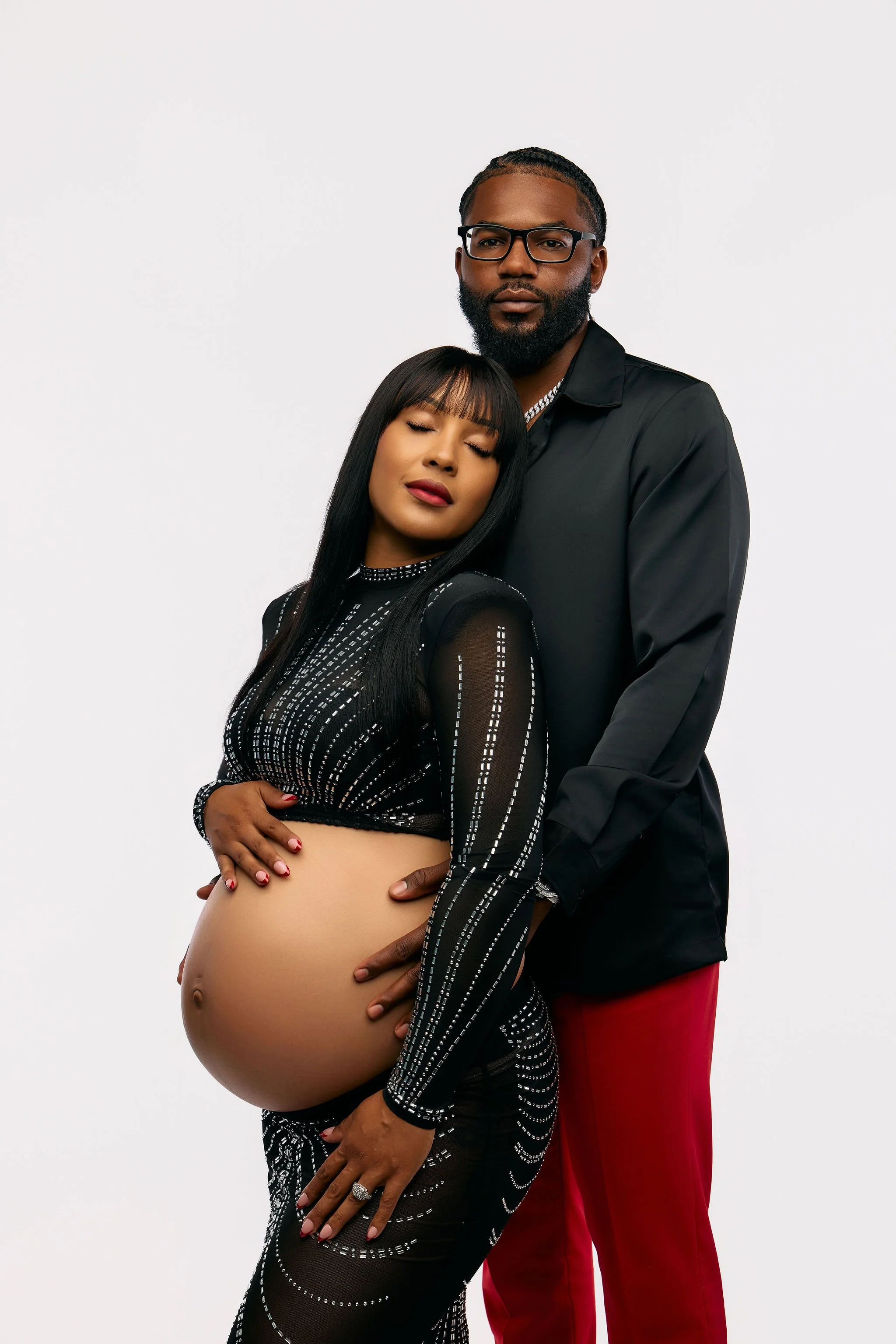 Expectant couple posing for a studio maternity portrait, with the mother cradling her baby bump in a black embellished gown and her partner standing behind her against a white background.