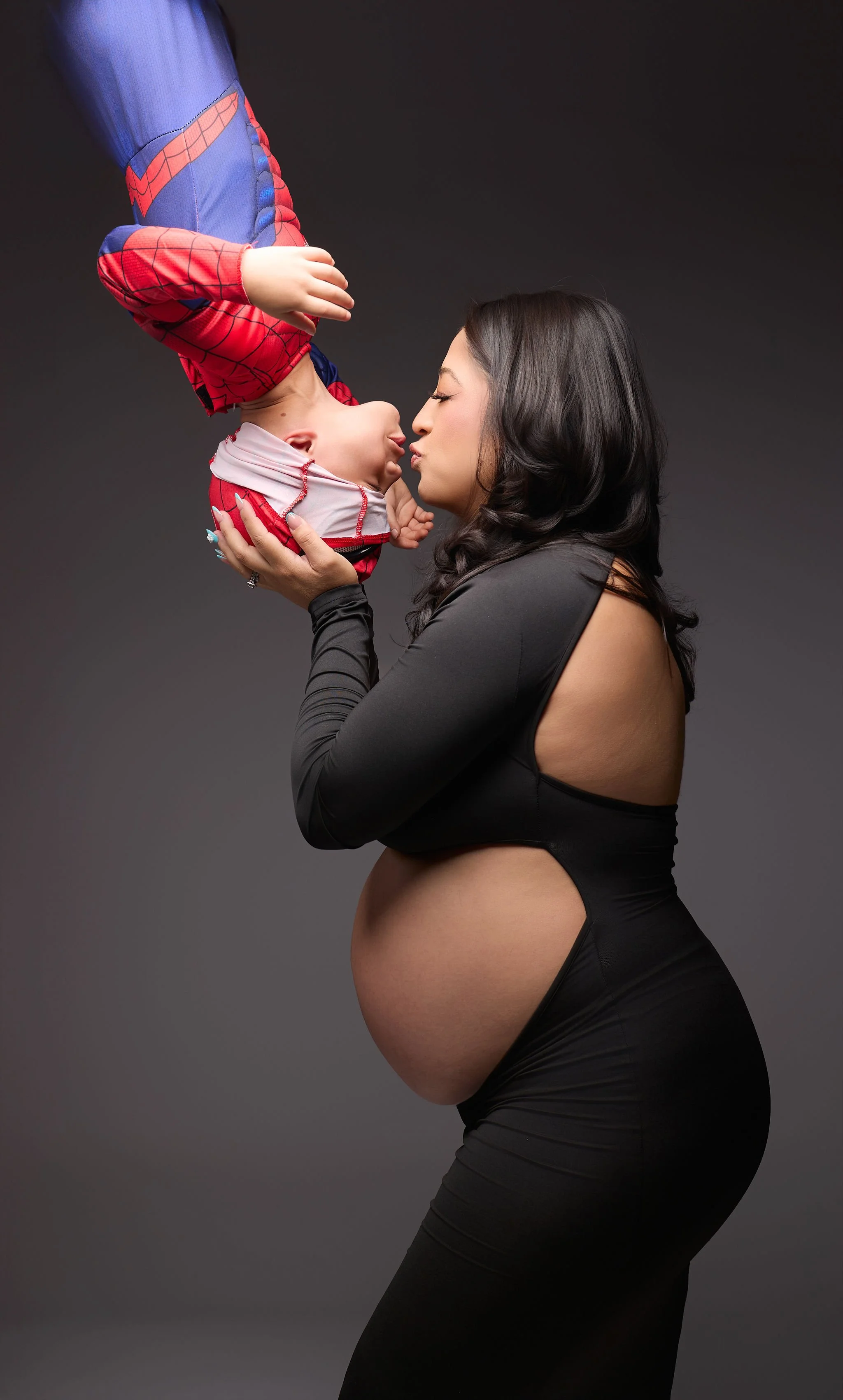 Pregnant woman in a black dress kissing a baby dressed in a Spider-Man costume against a dark gray studio background.