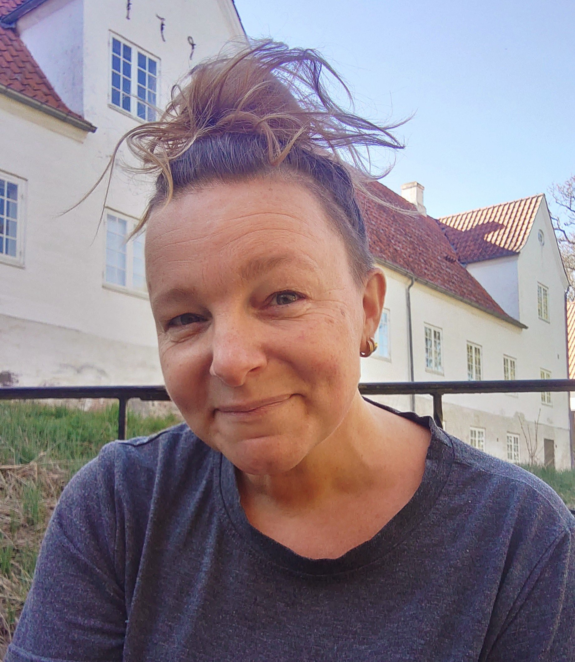 Person med kort, lysebrunt hår, der er ansigtet delvist skjult, iført en mørk, strikket sweater, står foran en hvid bygning med rødt tegltag, grønne områder og klar blå himmel.
