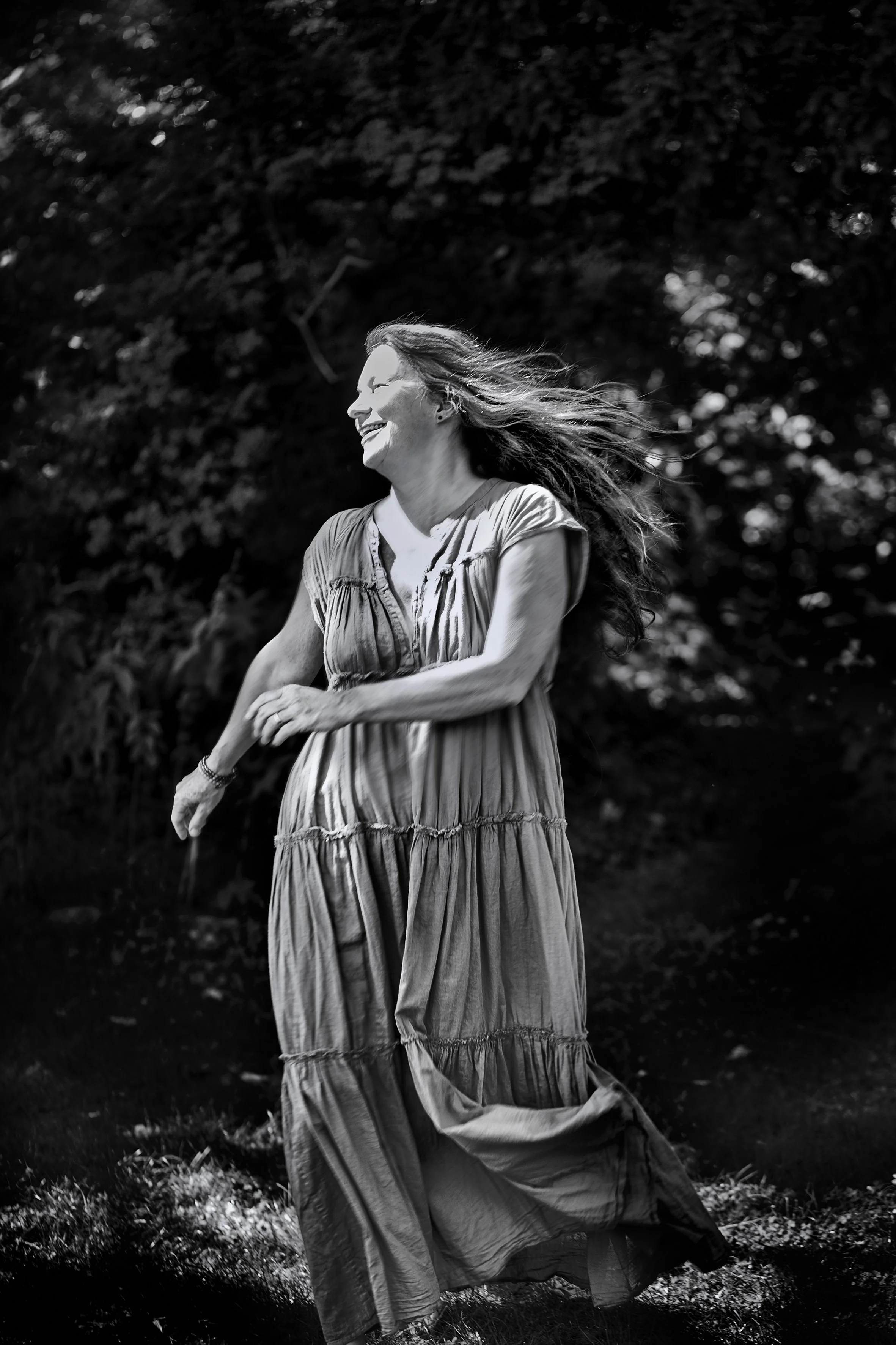 A woman in a long, striped dress smiles with closed eyes as she walks outdoors with the wind in her hair, surrounded by trees in the background.