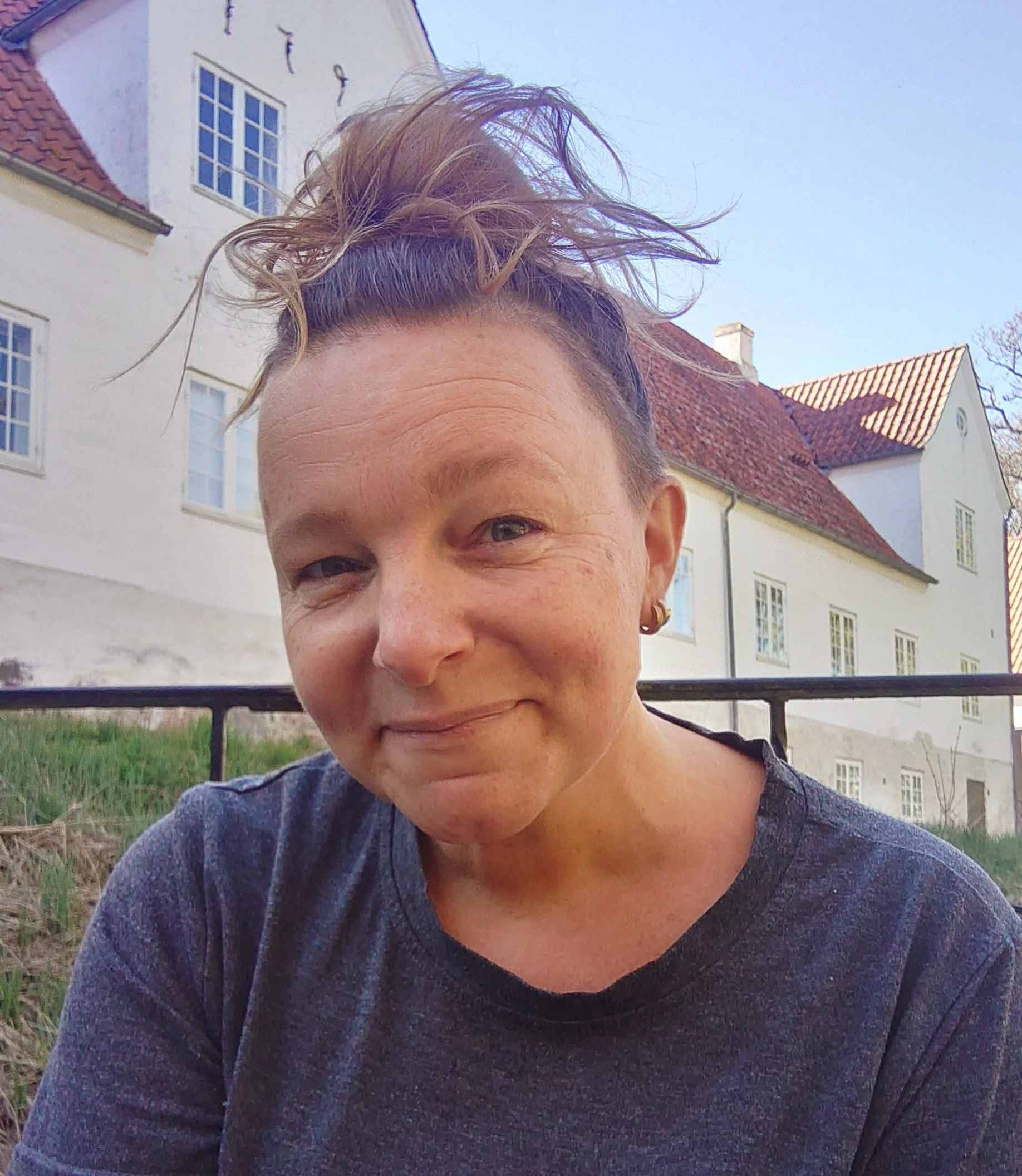 Person med lyst hår oppe i en løs knold, iført en mørk sweater, står foran en hvid bygning med rødt tegltag, mens det er klart vejr.
