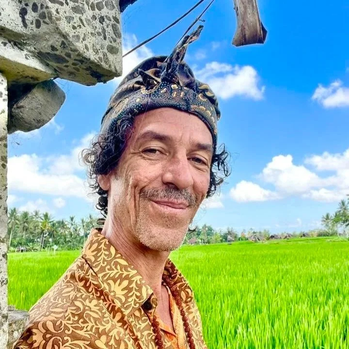 En mand med turban, smilende, foran grønne marker under blå himmel med skyer.