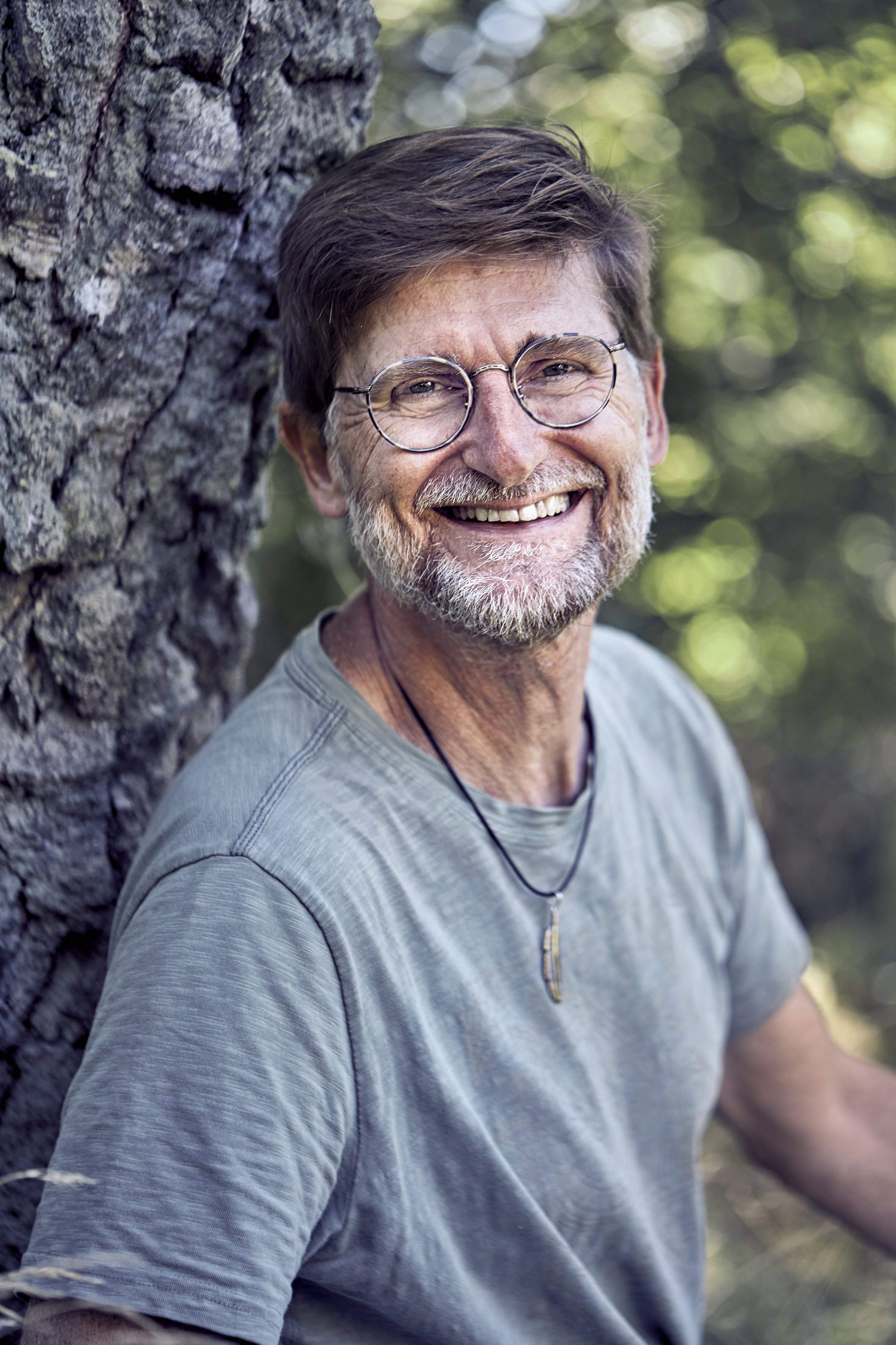 Et billede af en smilende ældre mand med brunt hår, briller og skæg, der sidder udendørs foran et træ, omgivet af grønne blade.