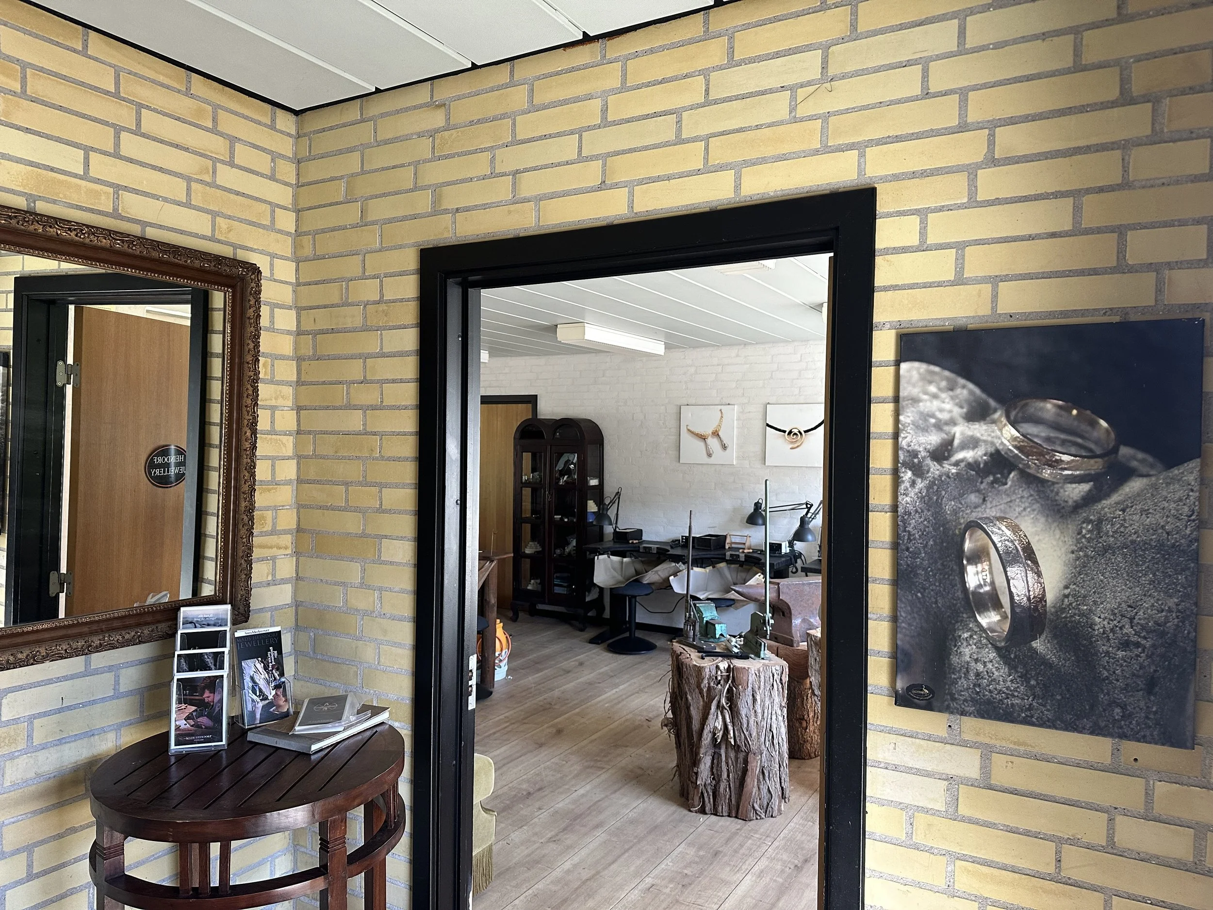 Indendørs butik med gule murstensvægge, store spejle, trappestige, træblok, og en åben dør, der viser et kontorområde med skriveborde og stole.