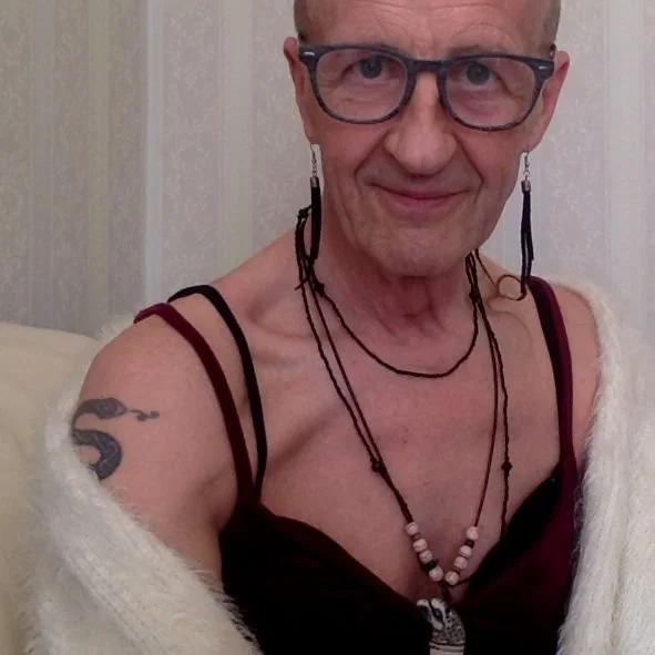 En person med glasses, halvnøgen med påklædning, halskæde, tattoo på armen, og en cardigan over skuldrene, siddende foran en væg.