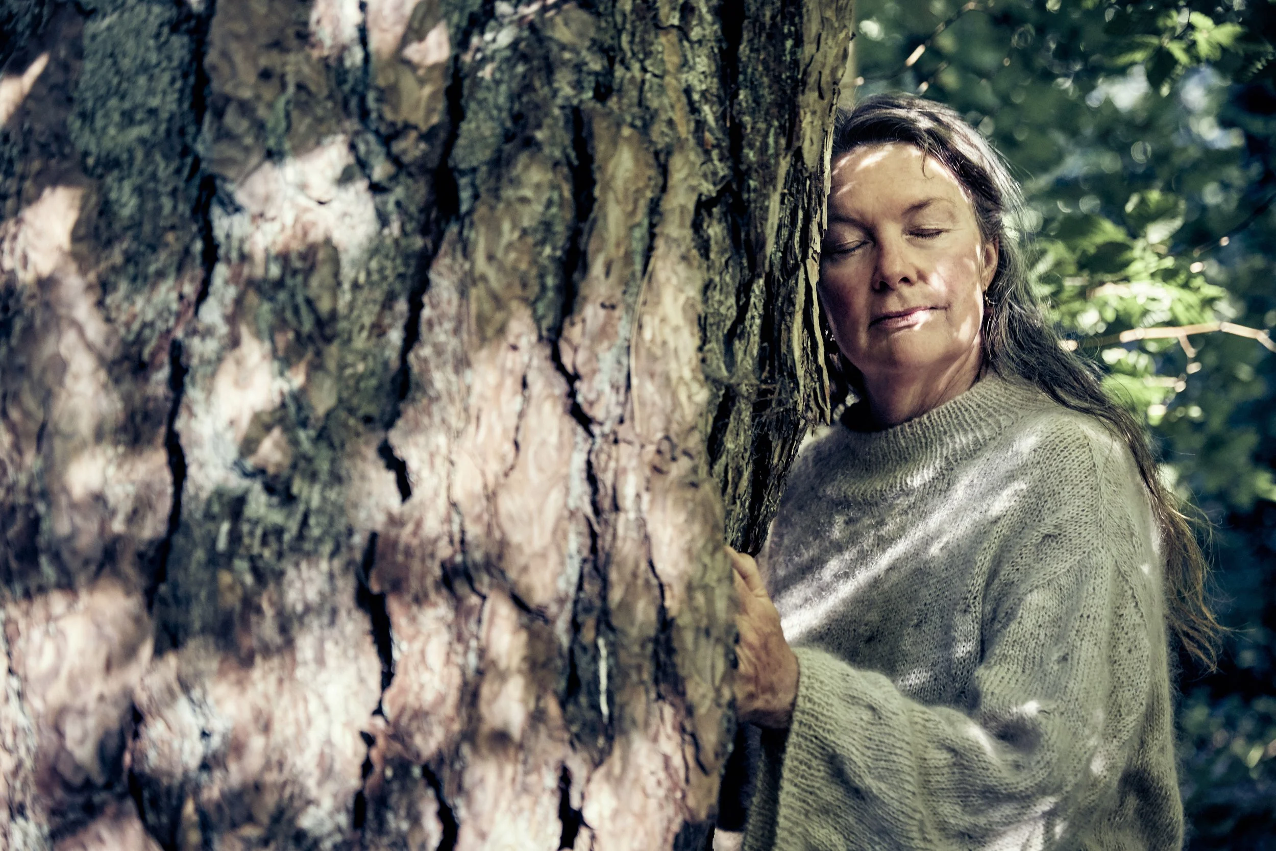 A woman with dark, wavy hair stands behind a tree, hugging it with closed eyes, surrounded by green leaves and forest.