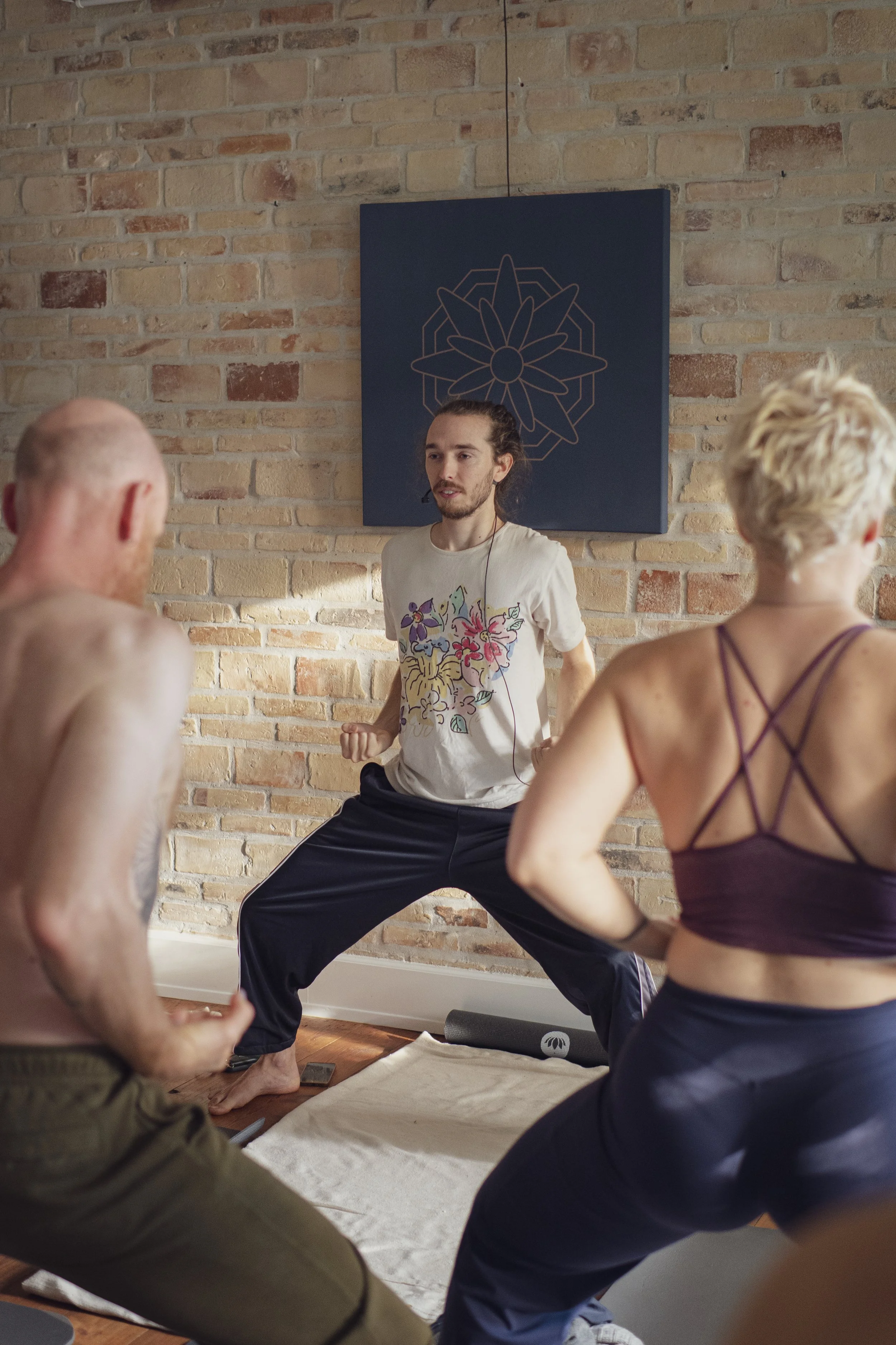 En yoga-instruktør står foran en gruppe mennesker, der udfører yoga i et rum med en murstensvæg. Instruktøren bærer en hvid t-shirt med farverige tegninger.