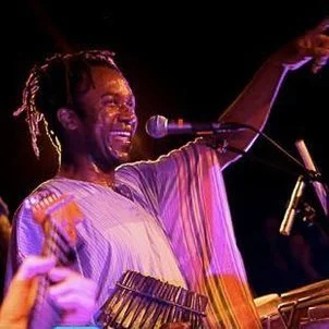 En glad musiker med dreadlocks, der spiller på et keyboard og synger i en mikrofon på scenen, med armene i vejret.