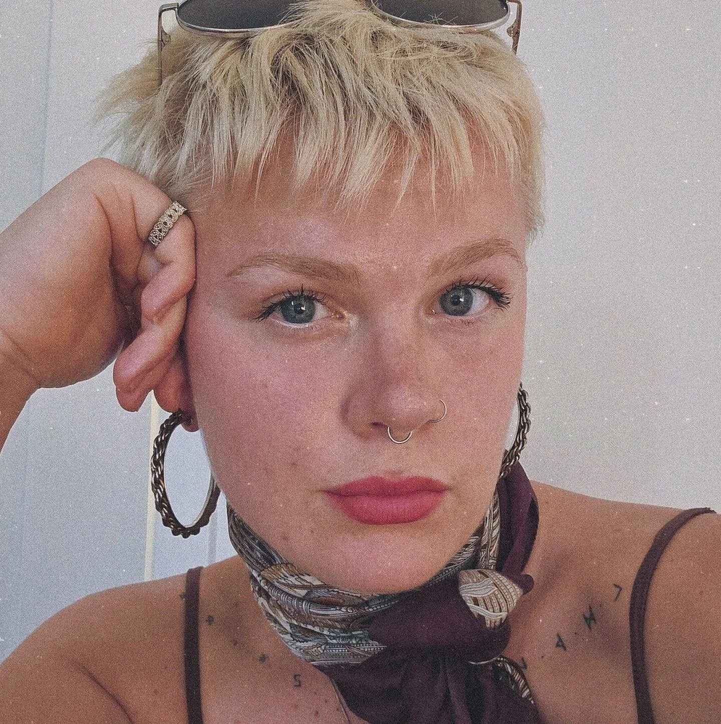 Et nærbillede af en ung kvinde med blond, kort, beskidt hår, der har på sig store øreringe, et næsepiercing, og en tatovering på skuldrene. Hun har på en scarf omkring halsen og bærer tynde stropper på tøjet.