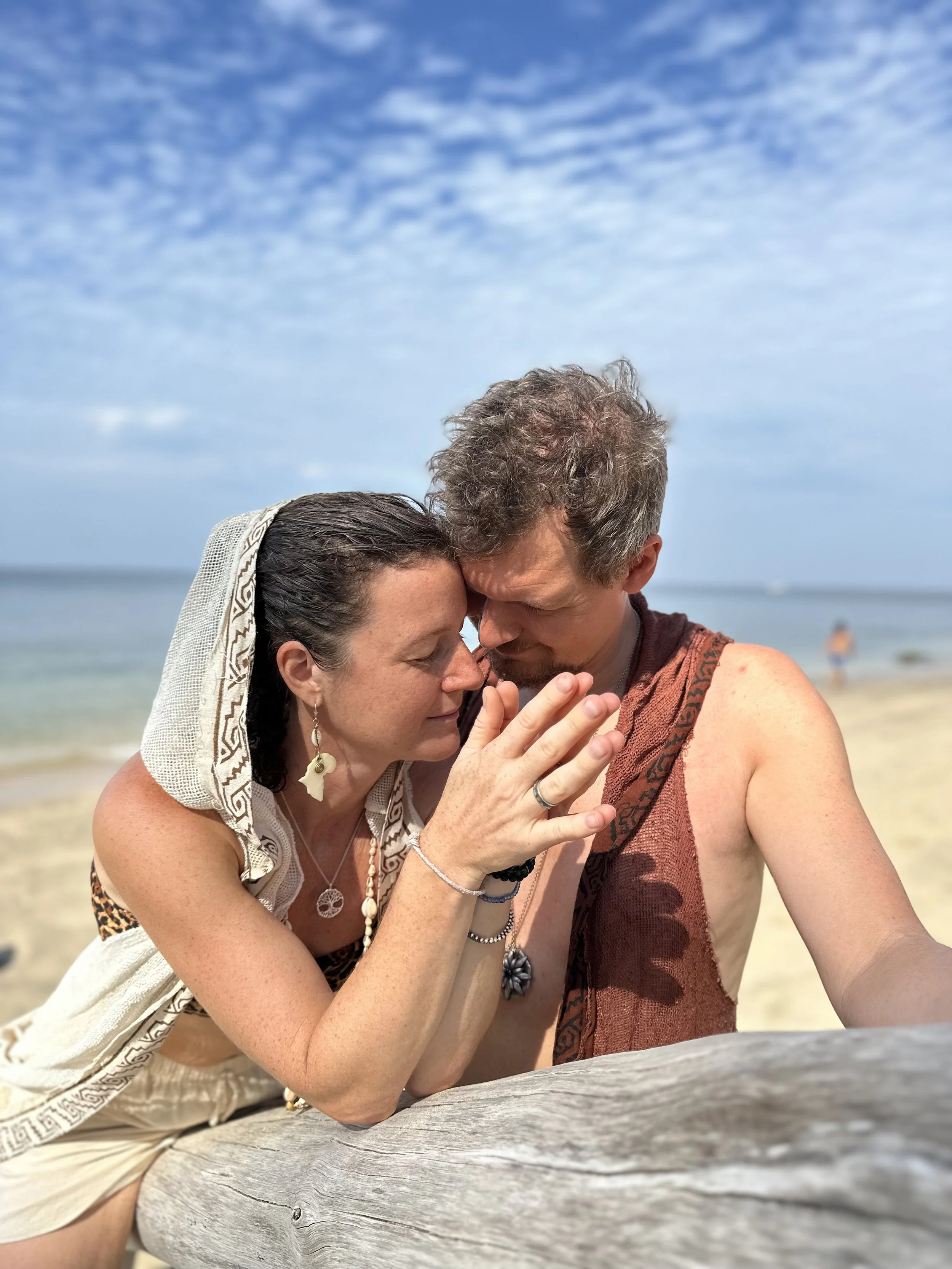 To mennesker, en kvinde og en mand, står tæt sammen med panderne og næserne mod hinanden på en strand; de holder hænder og har lukkede øjne, mens de deler et øjeblik af intimitet.