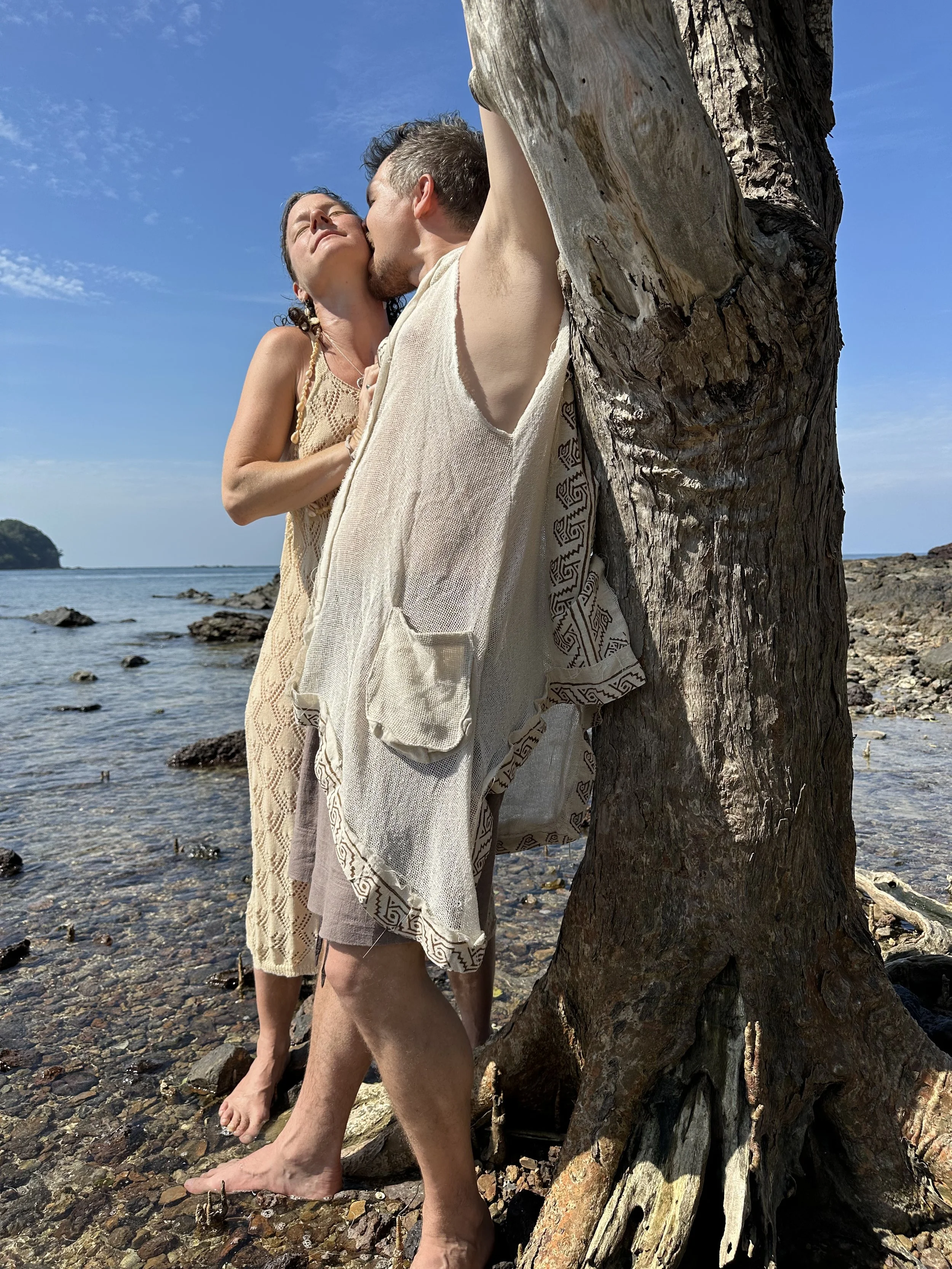To personer står tæt sammen ved en strand, den ene kysser den anden på kinden. Der er en stor træstamme i nærheden, og baggrund med hav og en lille ø.