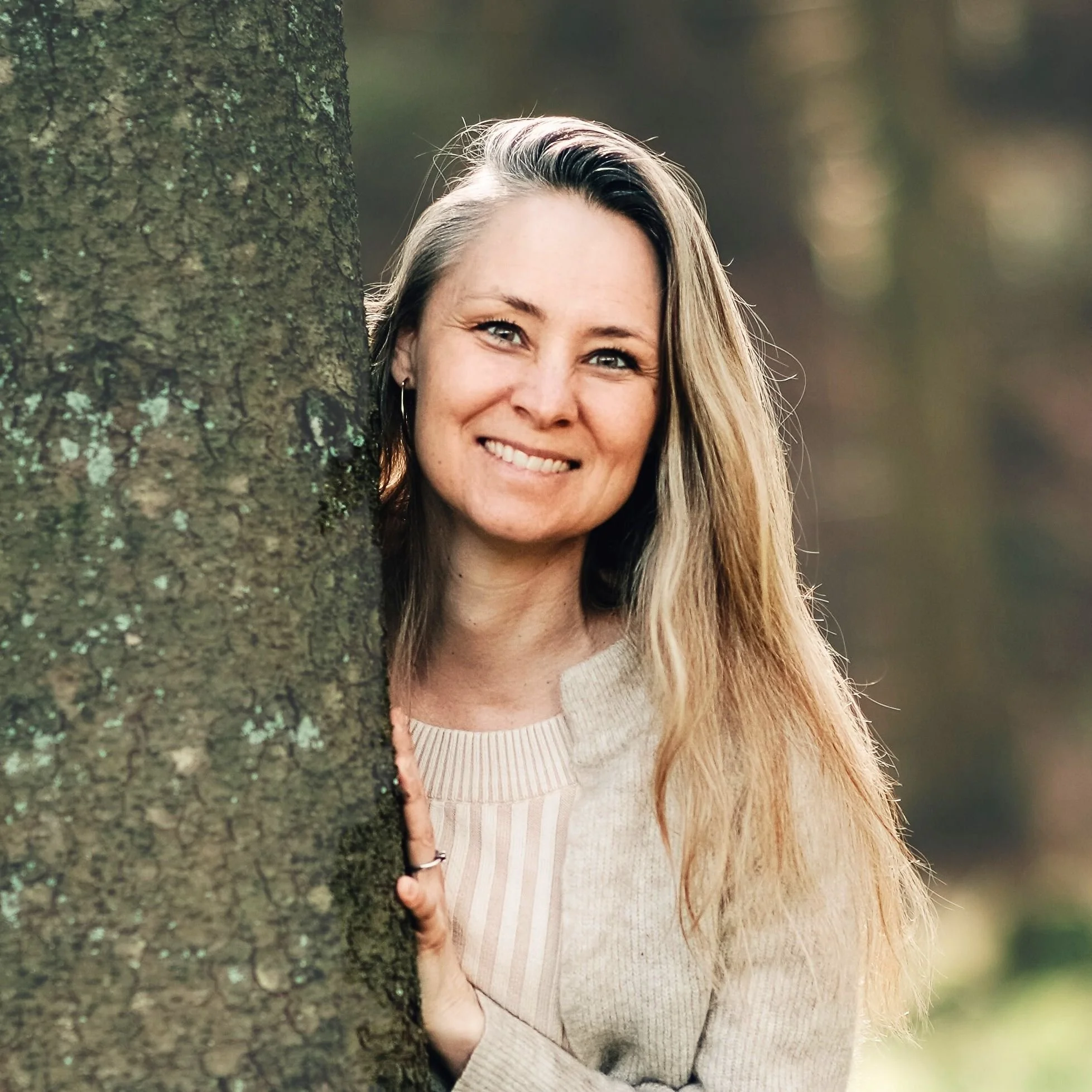 Smilende kvinde med langt blondt hår, iført beige sweater, står tæt ved et træ og smiler i en park.
