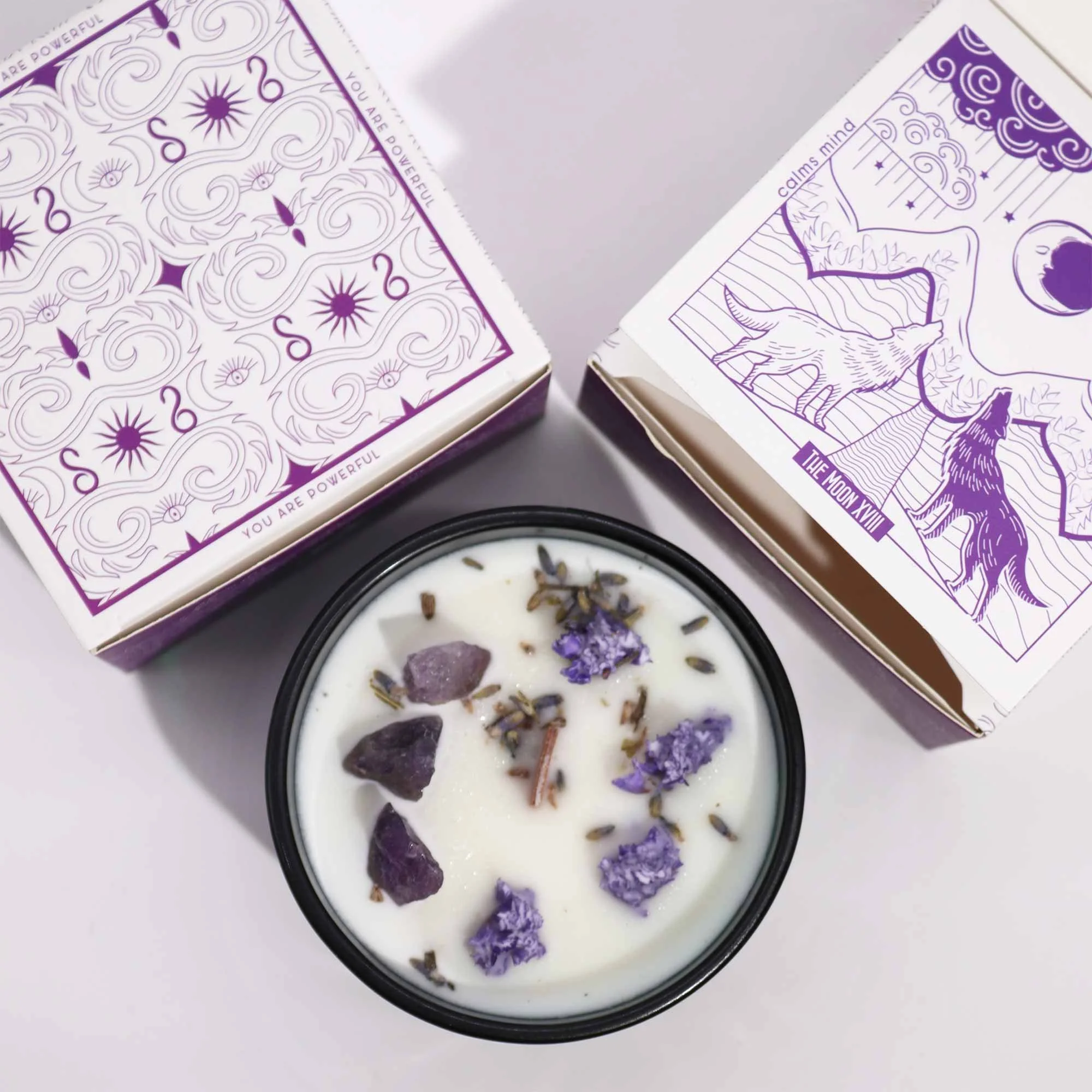 A lit white candle with purple stones, dried lavender, and purple flowers on top, placed on a white surface. Two decorated Tarot or oracle card boxes, one with celestial motifs and the other with wolf illustrations, are positioned above the candle.