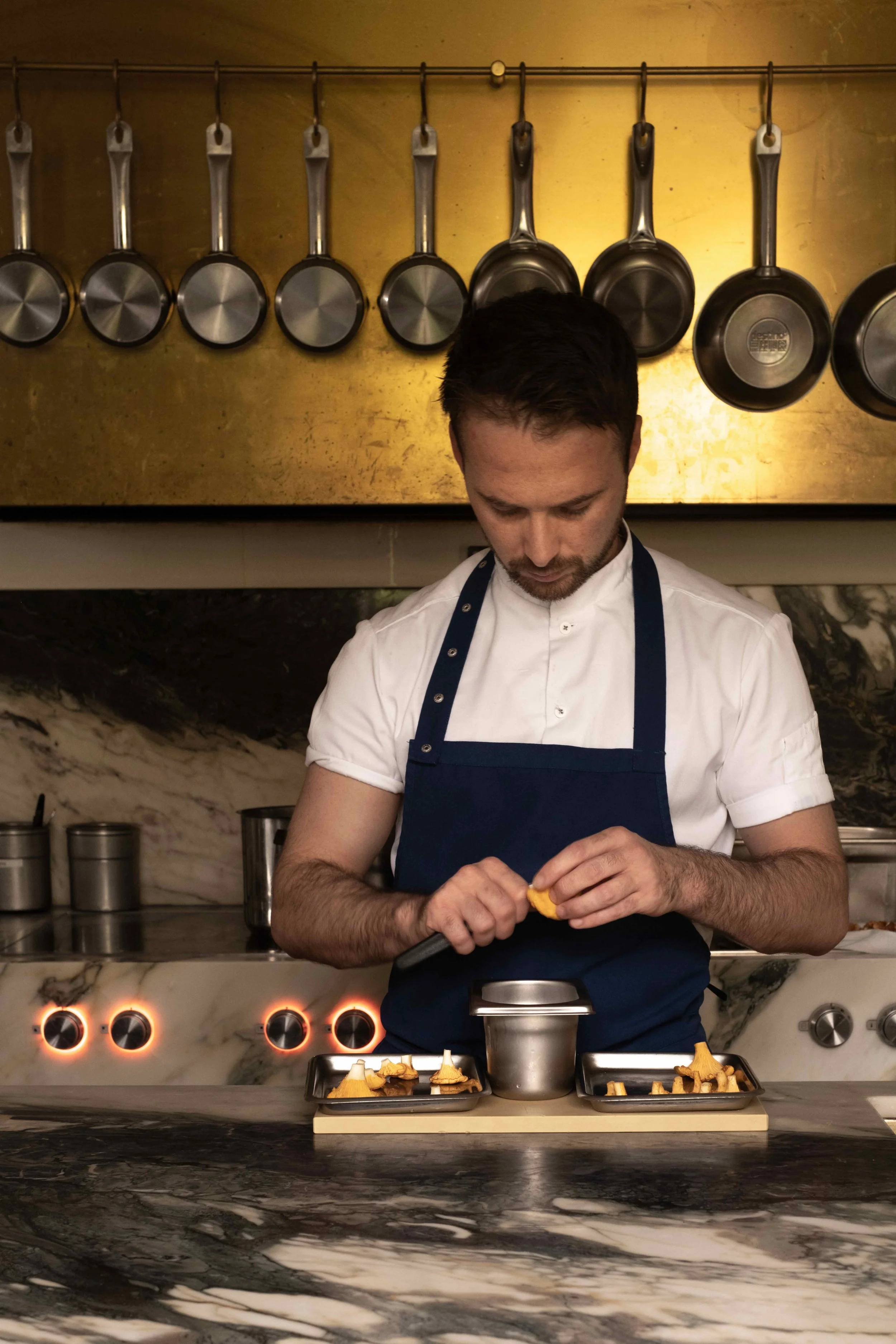 En mann i hvit kokkekjole og blå forkle som lager mat på en kjøkkenbenk av marmor, omgitt av gryter og panne, med flere panner hengt opp på en gullfarget vegg.