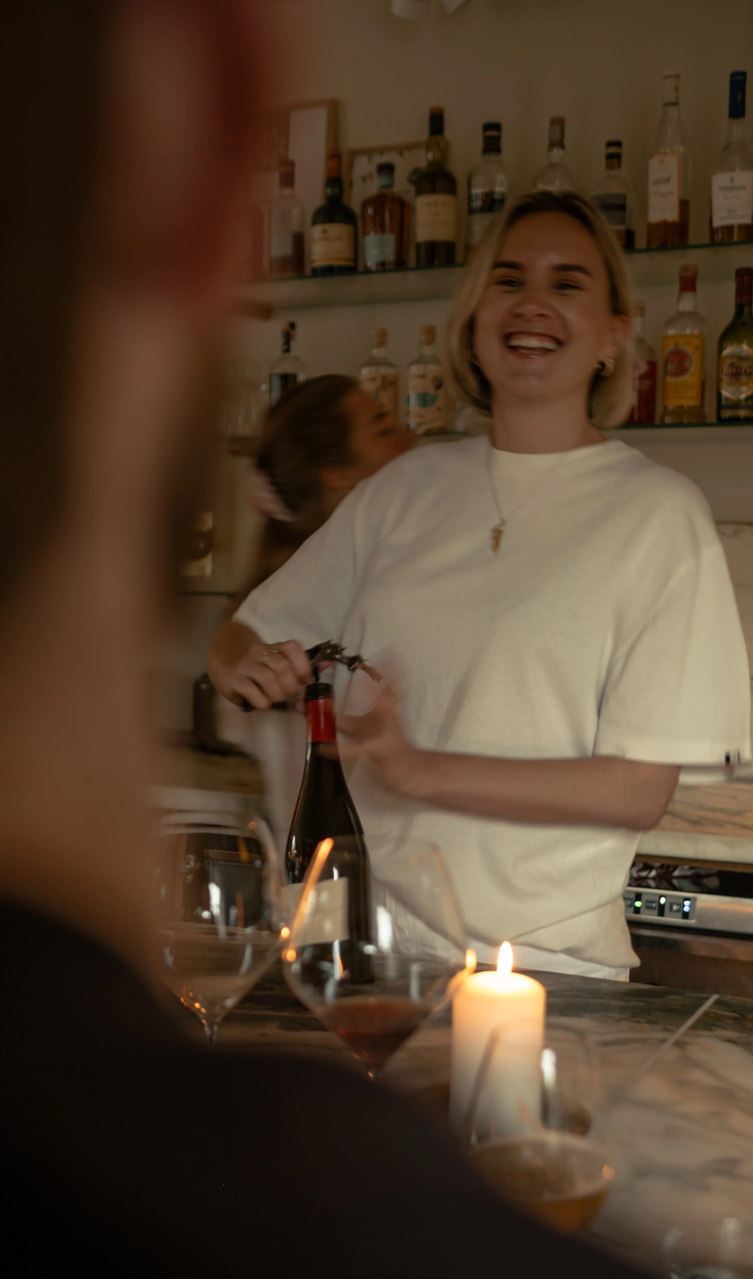 En kvinne med lys hår smiler og holder en korkekutter mens hun åpner en flaske vin i en bar, med flere vinflasker på hyller i bakgrunnen og levende lys på bordet.