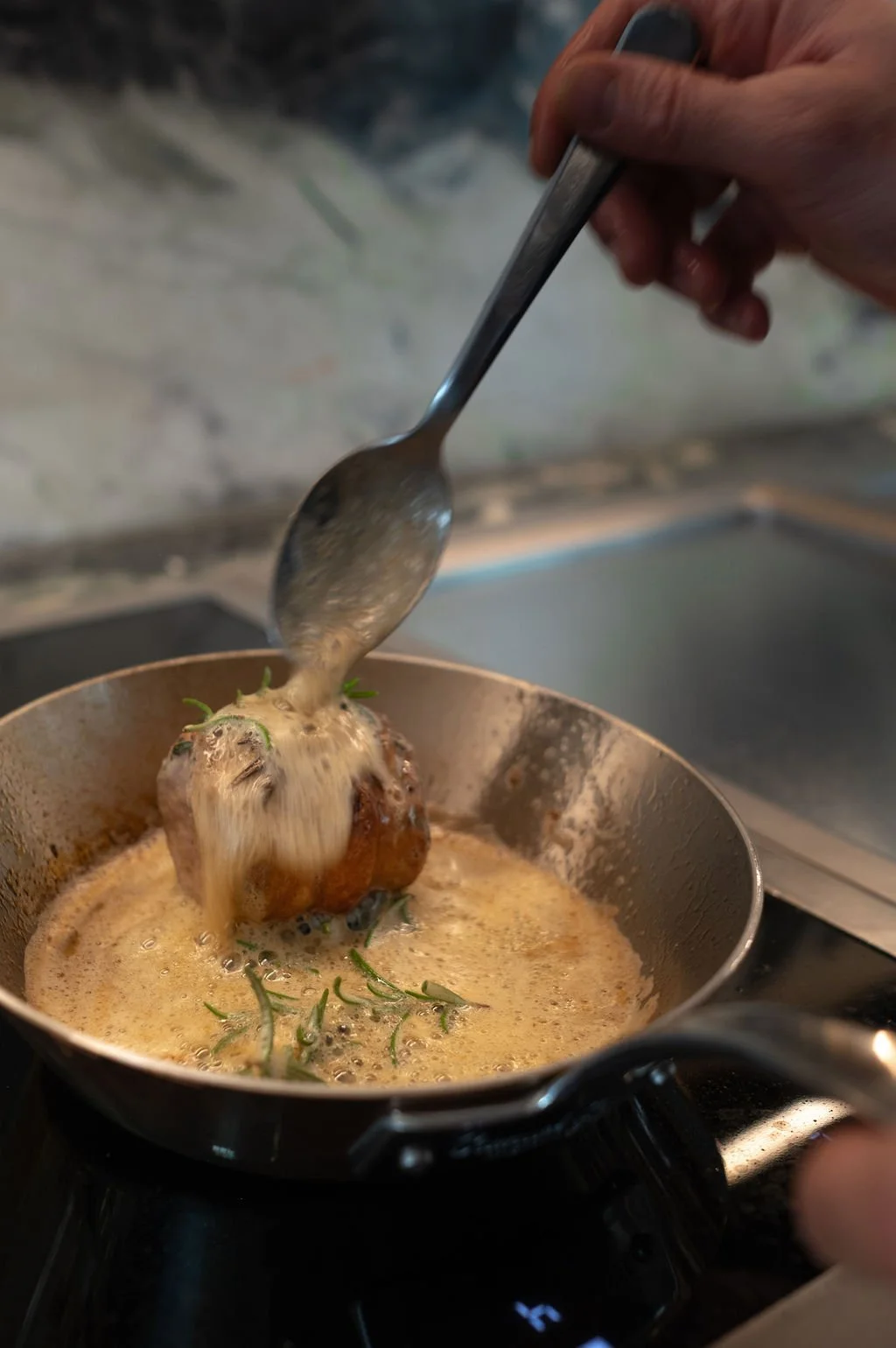 En person tilbereder en rett med en kjøttball i en panne med en saus, garnert med urter.