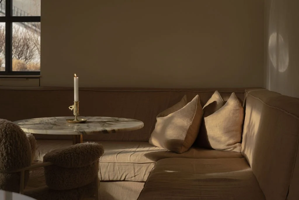 Et koselig hjørne med beige sofa, fire puter, et rundt bord med en lysestake og et vindu som slipper inn dagslys i et lyst rom.