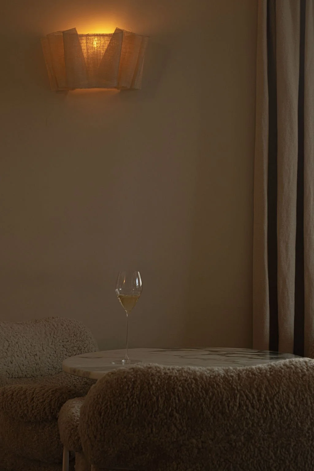 Et lyst rom med et bord, en flaske med vin, en vinflaske og en ustemplet krystallglass, ved siden av en sofa og gardiner, med en vegglampe som avgir varmt lys.