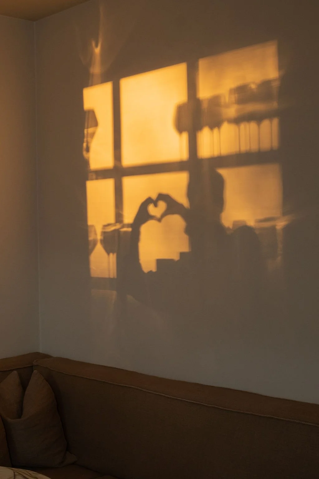 Skjult skygge av to personer som lager hjerteform med hendene foran et vindu med sollys som skinner gjennom, og skygger av bøker på hylle som er synlige i bakgrunnen.