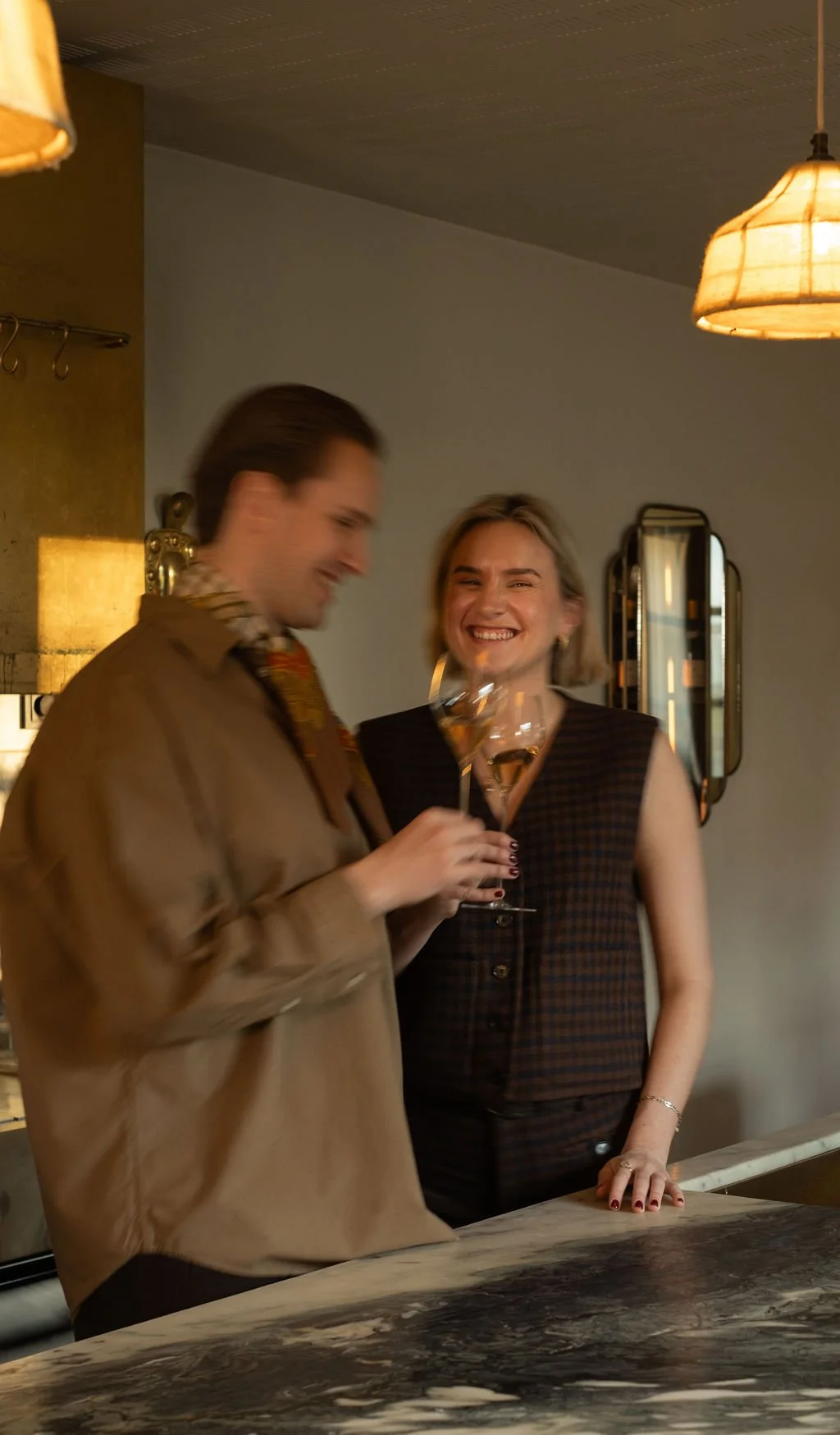 To personer ler og skåler med glass vin i en innendørs setting, hyggelig atmosfære.