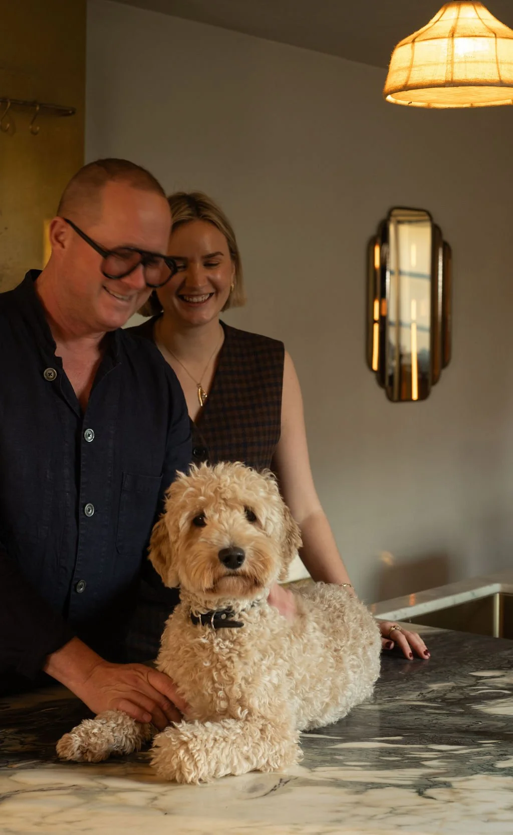 To voksne personer, en mann og en kvinne, som ler og er glade med en liten krøllete hund på et bord inne i et rom med moderne dekorasjoner og varm belysning.