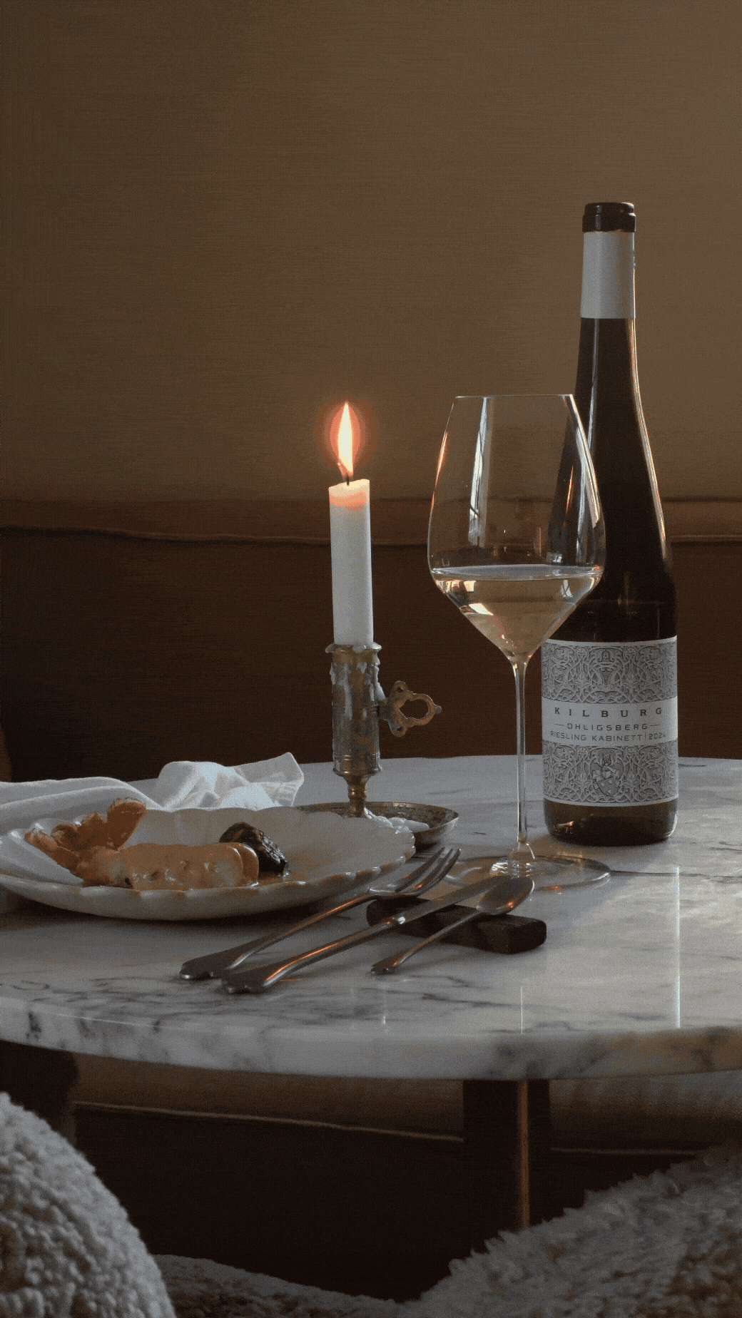 Et bord med et tørketroskudd av lys, et vinglass med hvitvin, en vinflaske, en tallerken med mat, og bestikk, satt opp på et marmorbord i et rom med dempet lys.