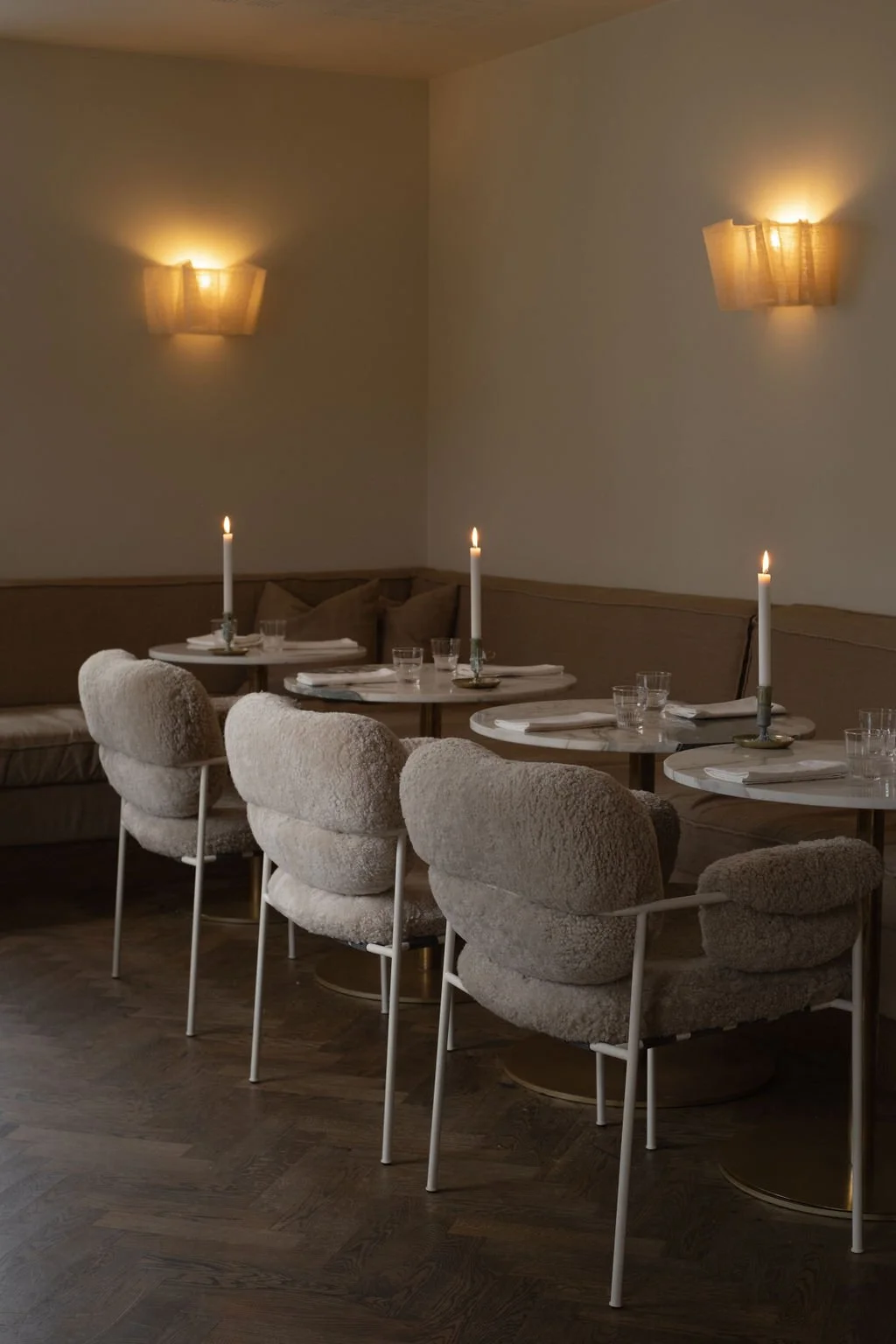 Elegant restaurant med fire bord, hver med krystallvaser og tente lys, komfortable stoler med ullstoff, minimalistisk vegglys, og tremøbler.