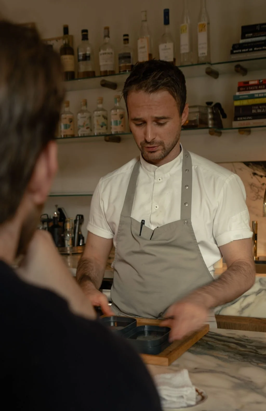En mann som er kledd i hvit kokkjakke og grå forkle, står bak en bar eller arbeidsbenk, og ser ned med en bekymret eller frustrert uttrykk. I bakgrunnen er det hyller med flasker og kokebøker.