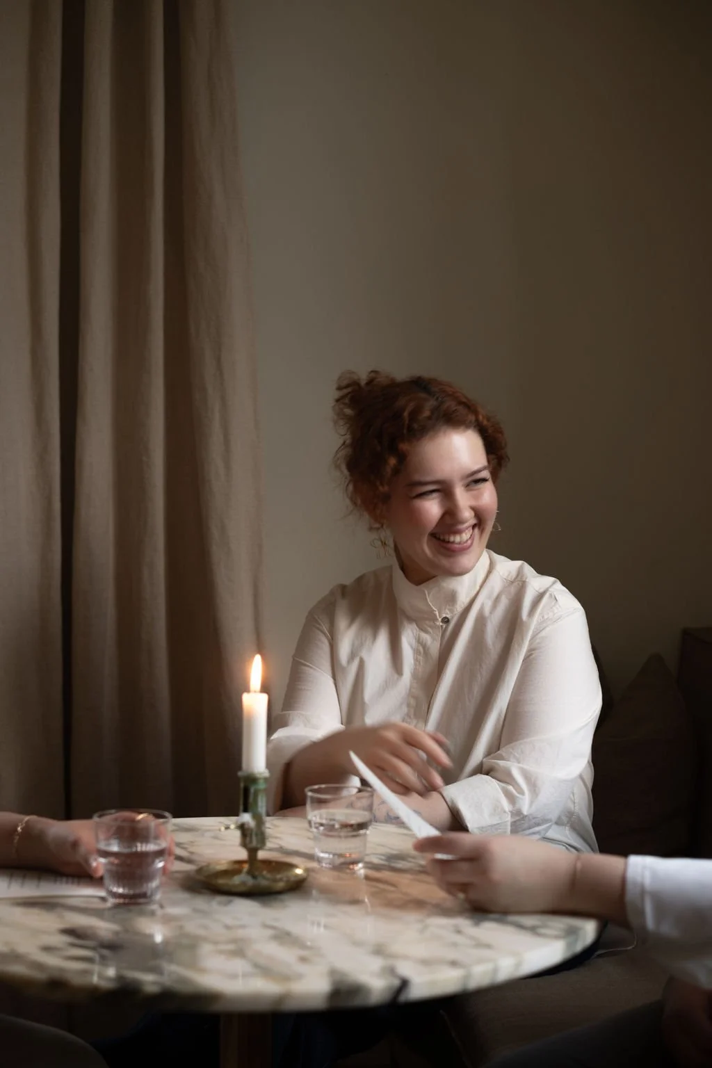 En kvinne med krøllete, brunt hår som smiler, sitter ved et bord med en lysende stearinlys, to glass med vann og en person som gir henne en serviett.
