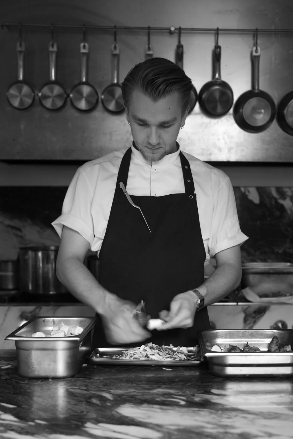 En mann i kokkepynt med svart forkle og hvit kokkeklær, som skjærer grønnsaker i et kjøkken.