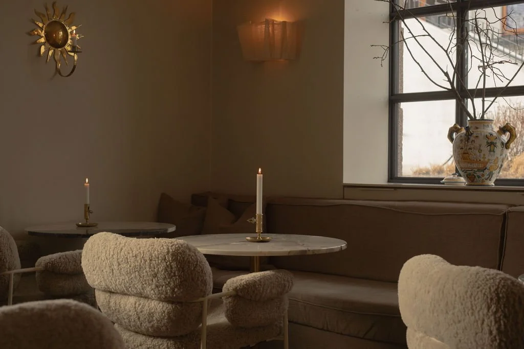 Innendørs stue med beige møbler, to stearinlys på bordene, stor vase med blomster på vinduskarmen, og et vindu med uteseende av vinterlandskap utenfor.