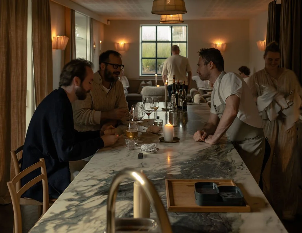 Innendørs restaurantsetting med flere personer, noen som sitter og snakker, og en servitør som står ved baren.