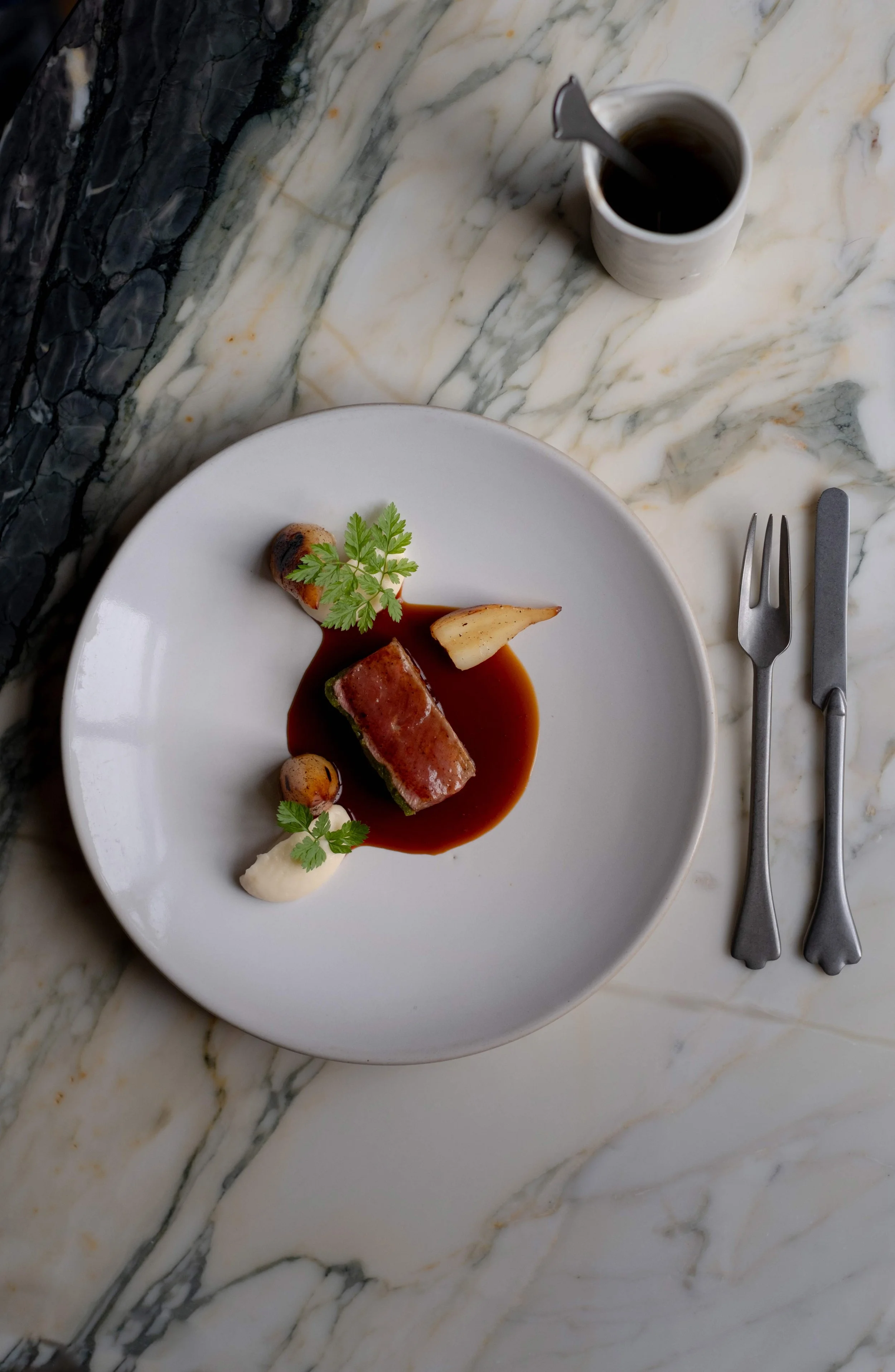 Elegant tallerken med kjøttstykke i saus, garnityr av urter, poteter og grønnsaker, ved siden av en kopp kaffe. Sølvbestikk ligger til høyre. Marmerbord under.