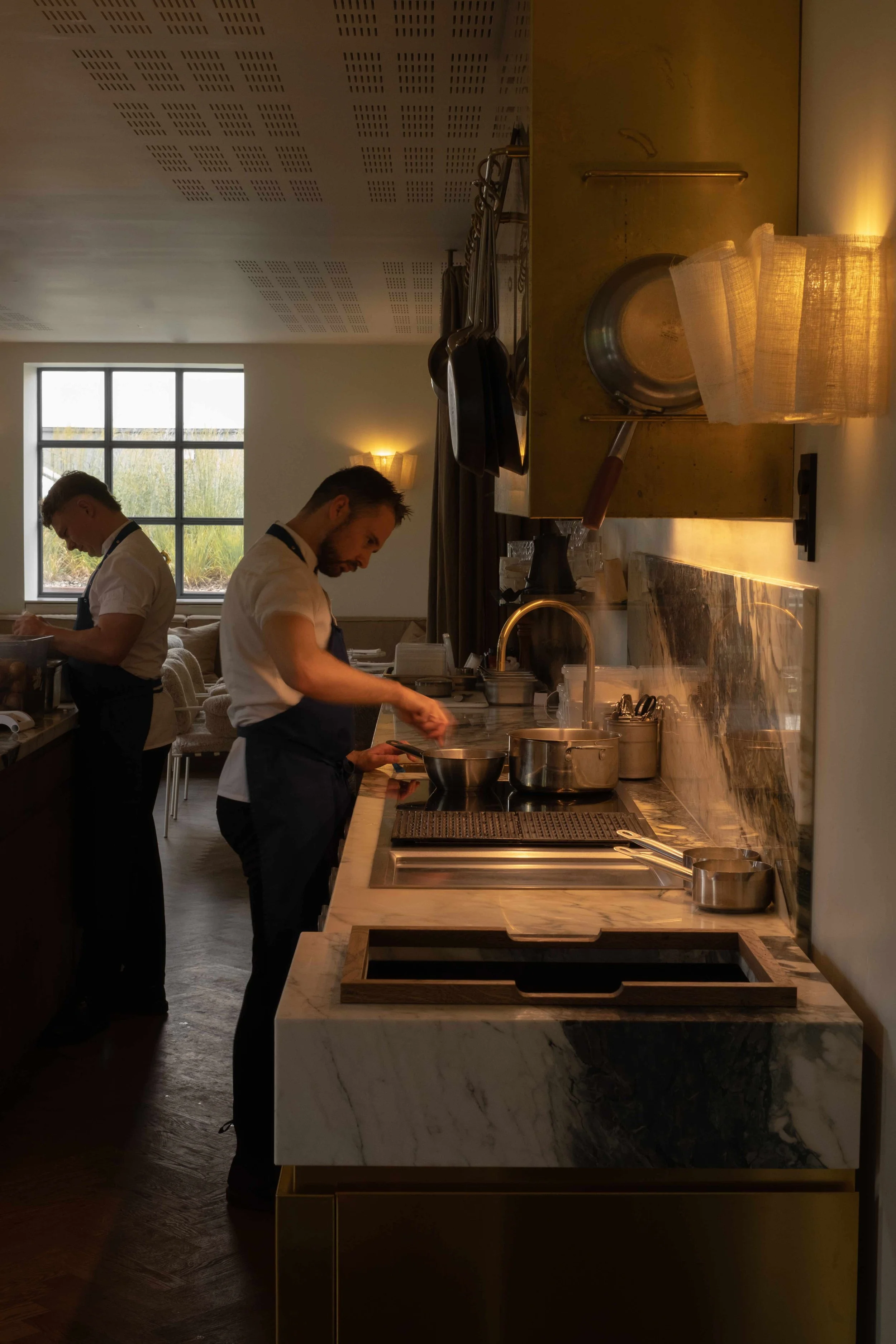 To kokker i en koselig restaurantkjøkken, som forbereder mat, med et vindu og varm belysning.