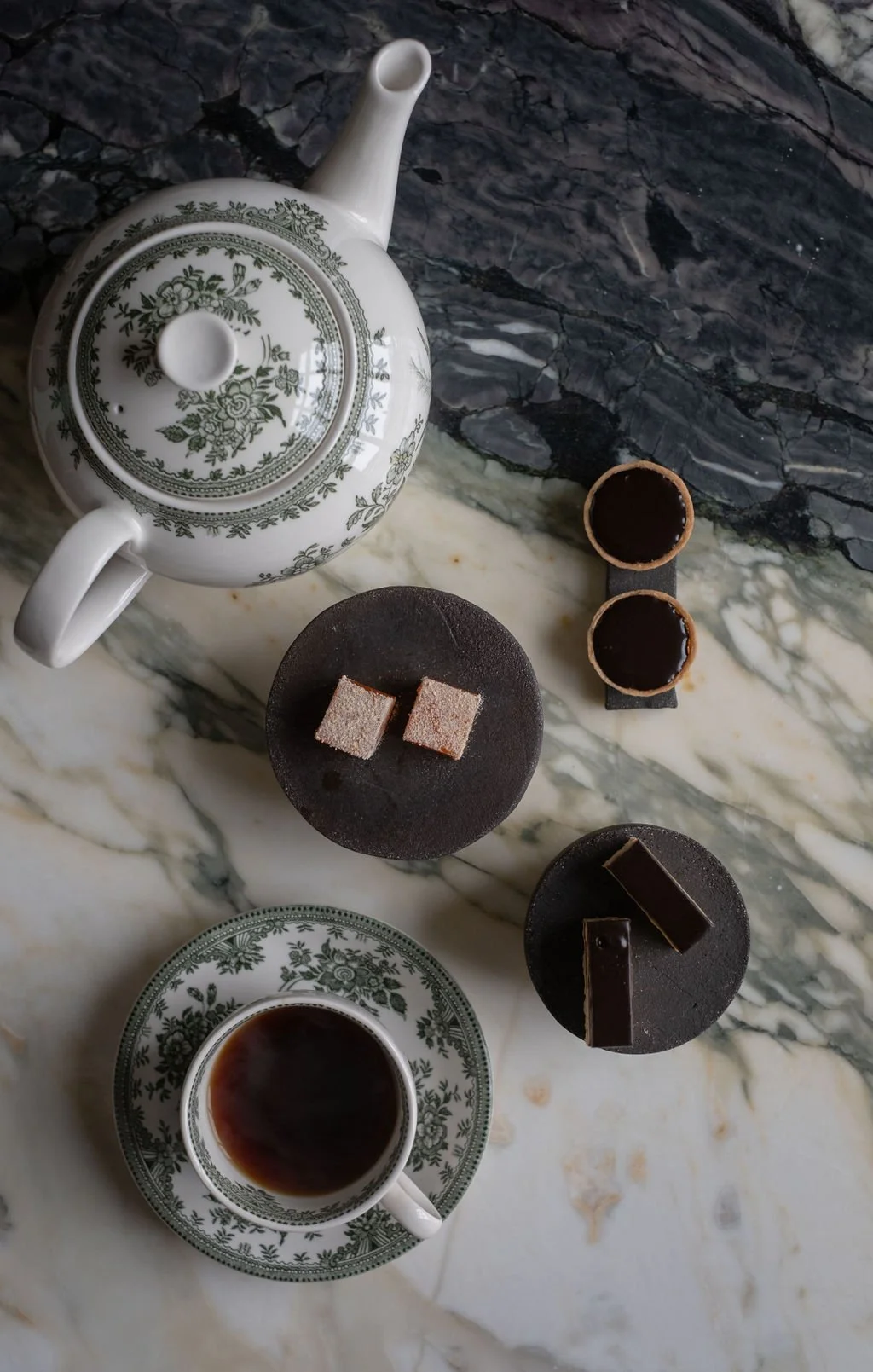 En kopp med svart kaffe, en hvit tekanne med grønt mønster, tre små sjokoladekaker på svarte plater, og to mini sjokoladetuber på svart brett på en marmorbord.