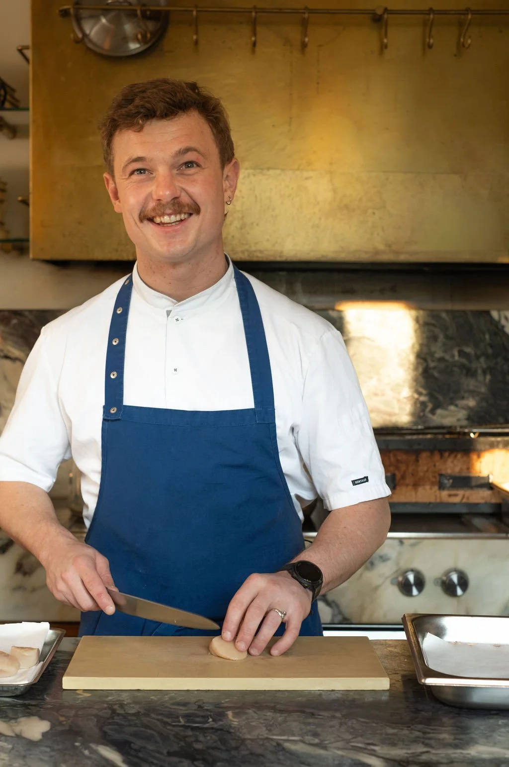 En mann med bart i hvit kokkegenser og blå forkle som skjærer en prosjonl. Han smiler og står i et kjøkken.