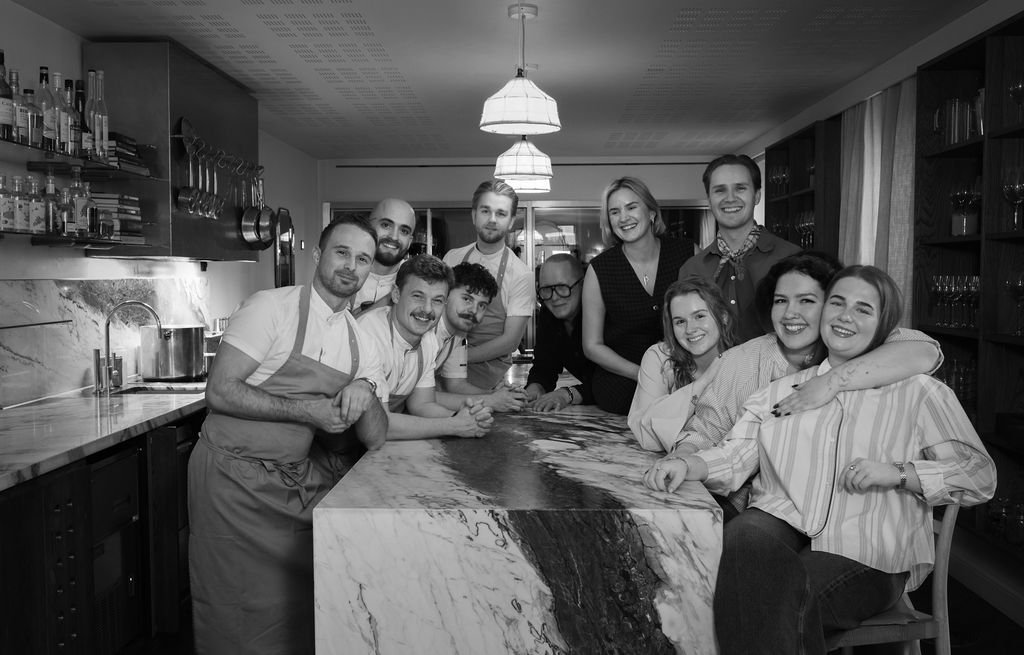 En gruppe mennesker i et kjøkken, noen som smiler og poserer for et bilde. Flere av dem er kledd i kjøkkenuniformer.