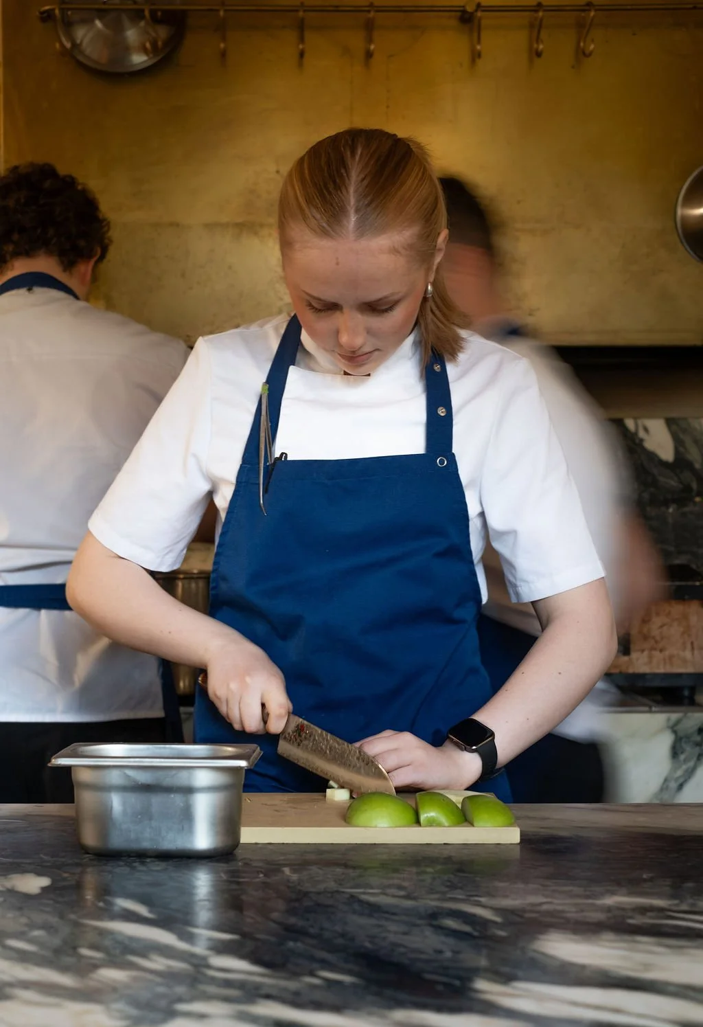 Kvinne i kokkeforkle skjærer grønne epler på et skjærebrett i et kjøkken.