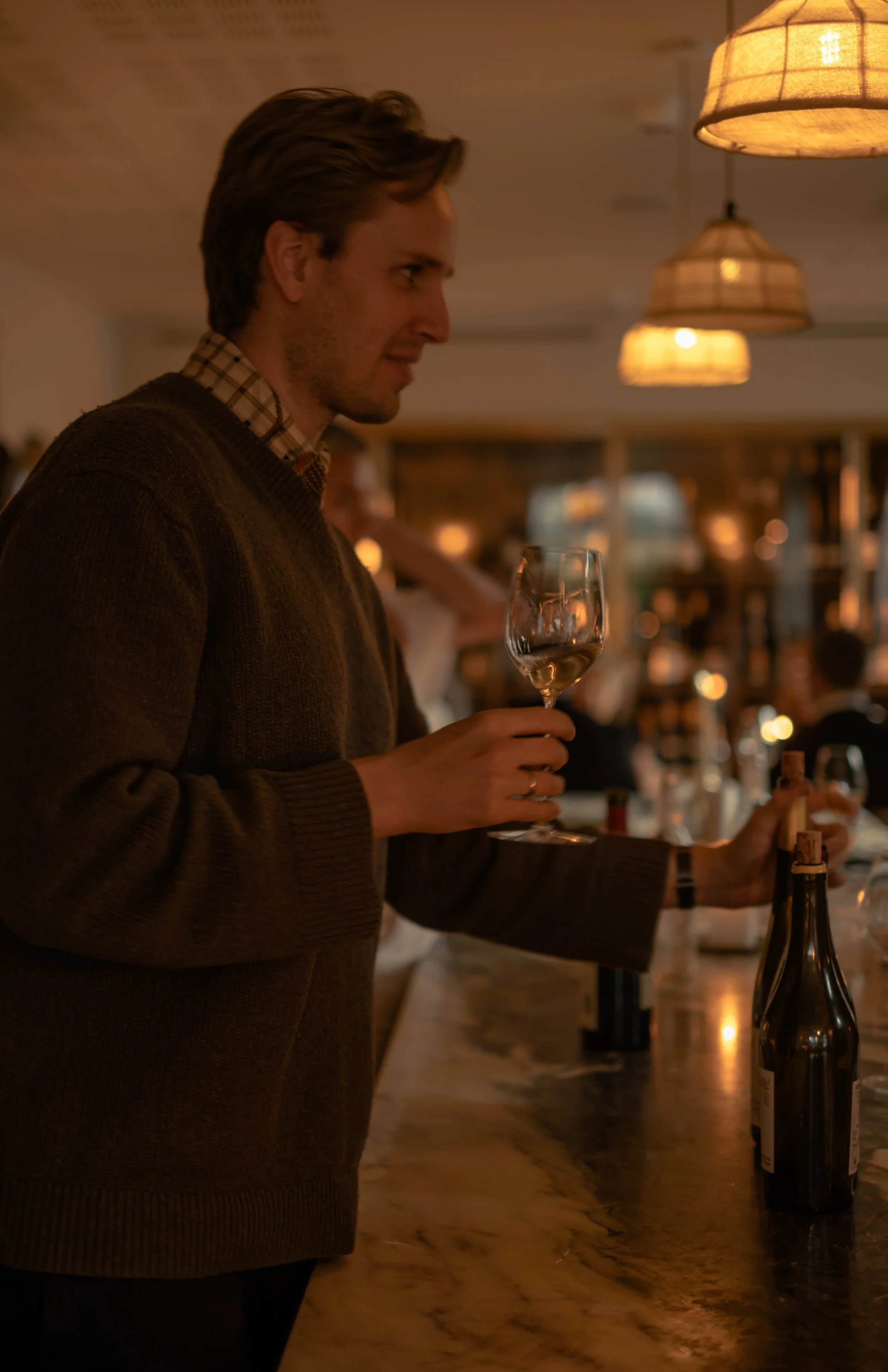 En mann med brunt genser og rutete skjorte holder et vinglass med hvitvin på en bar eller restaurant med varm belysning.