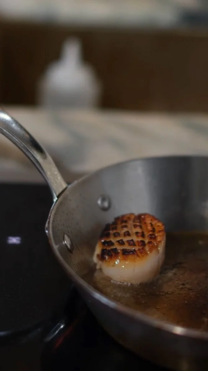 Sølv stekepanne med grillet scampi i olje, på koketopp. Bakgrunn med sølvflaske.