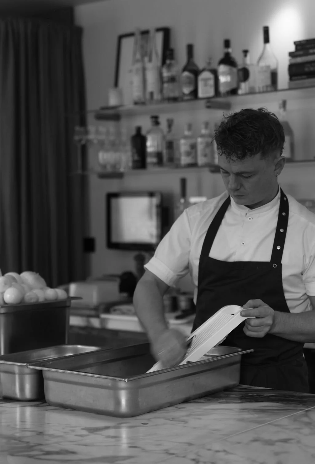 En mann iført en hvit skjorte og svart forkle står på kjøkkenet, og sprayer innhold i en metallboks, med en rekke kokebøker og flasker på hyller i bakgrunnen.