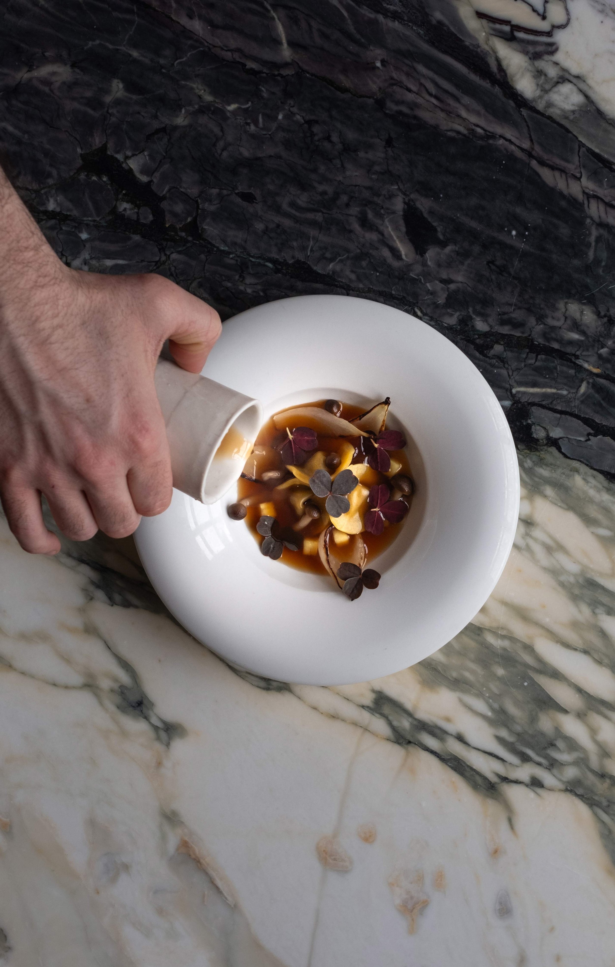 En hånd helle en brun saus eller væske over en tallerken med små matvarer og urter, plassert på en hvit tallerken på en marmorbord.