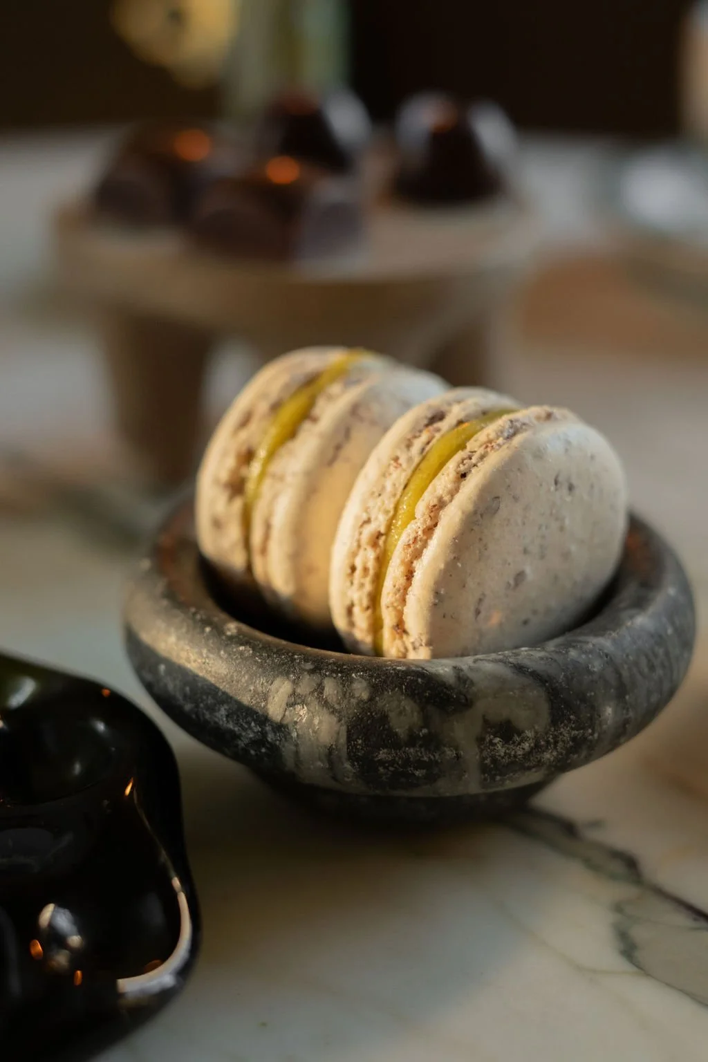 Macarons i en steinur skål med flere sjokolade- og vaniljemacarons innenfor. I bakgrunnen er det en tallerken med små sjokoladekaker.